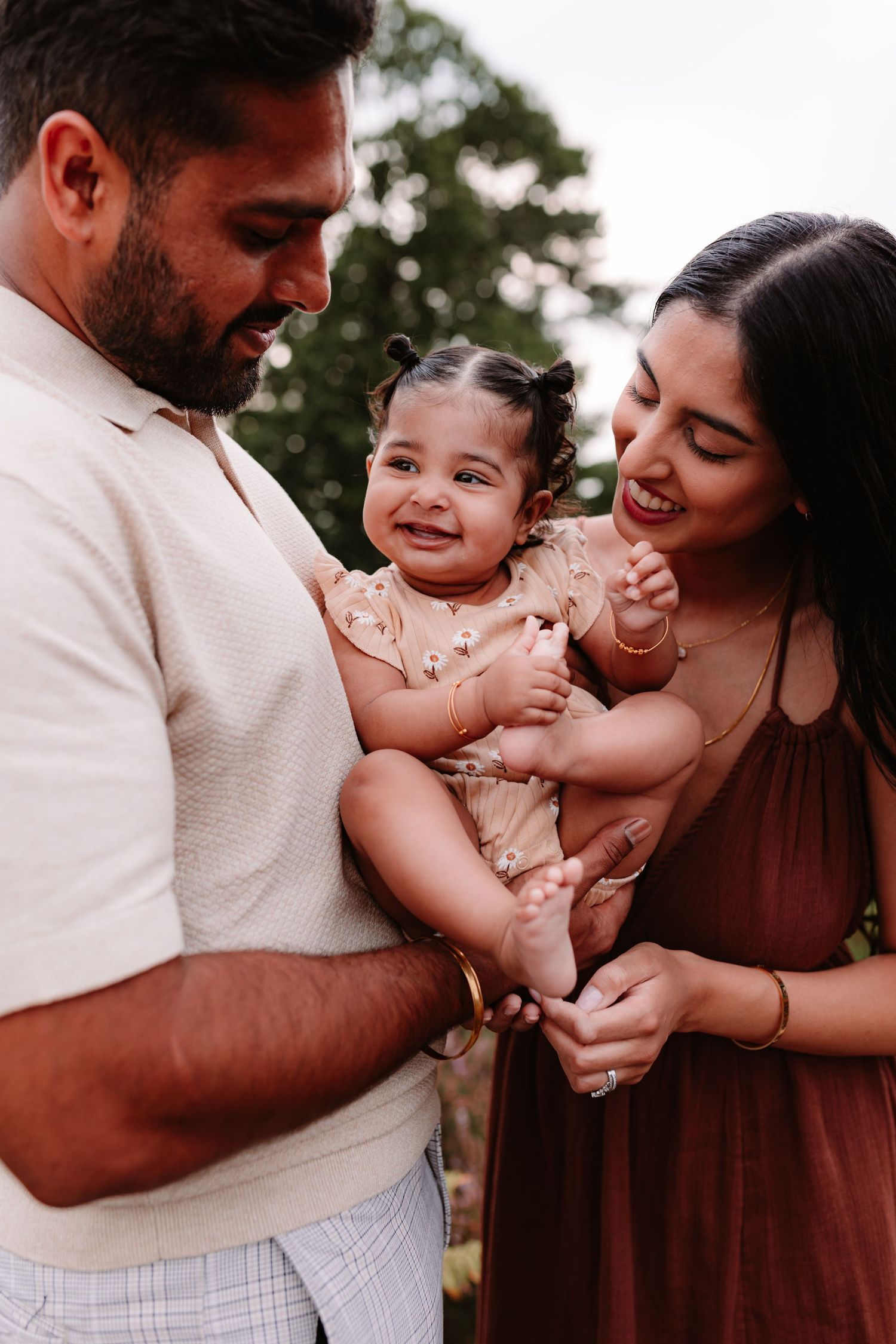 Happy family portrait with parents holding their smiling baby girl outdoors in a garden setting.