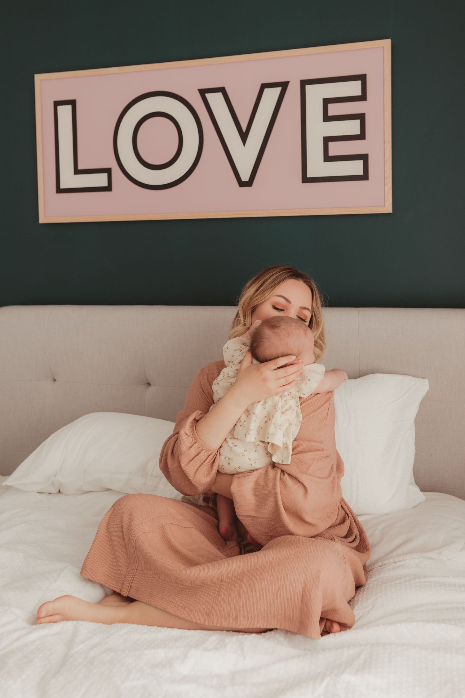 Blonde woman in pink pajamas sitting cross-legged on bed cuddling newborn baby under a LOVE sign.