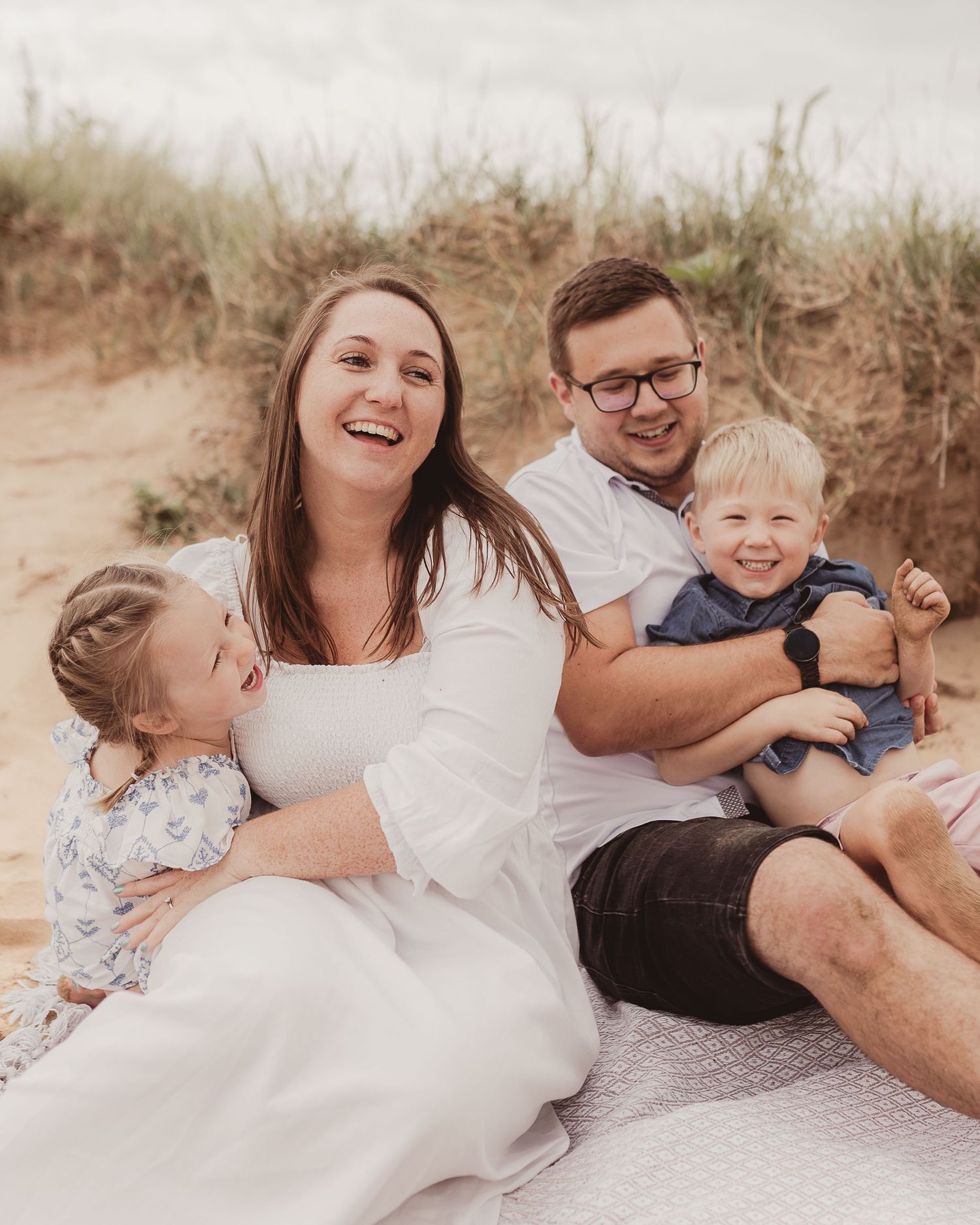 Laughing family of four relaxing together on a blanket at a sandy beach on an overcast day.