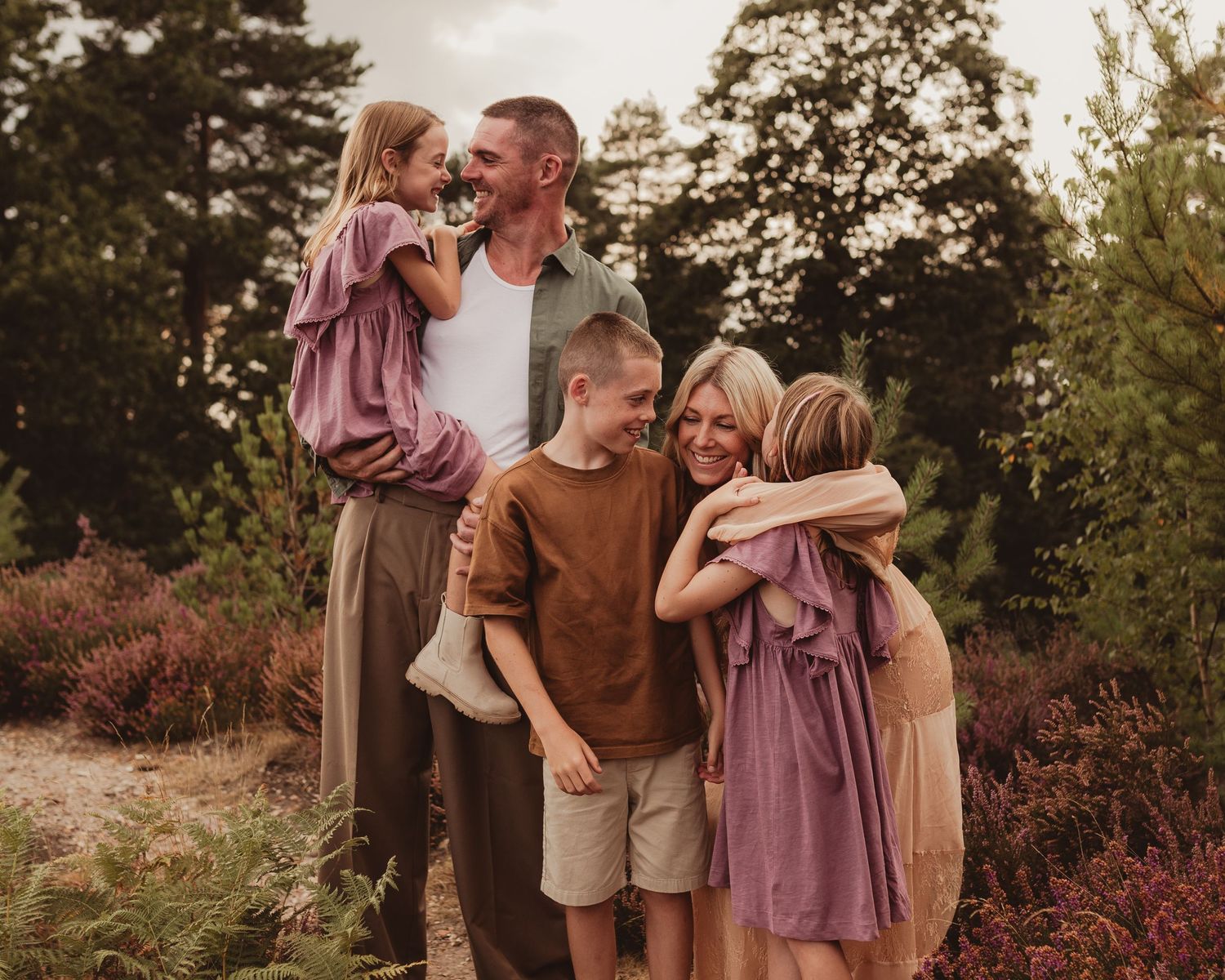 Happy family of five sharing hugs and smiles outdoors surrounded by lush greenery and purple heather.