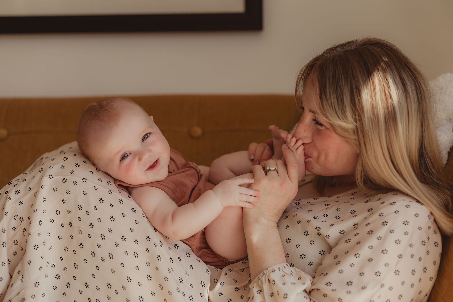 A mother tenderly kisses her smiling baby's feet while relaxing together on a cozy bed with soft neutral bedding.