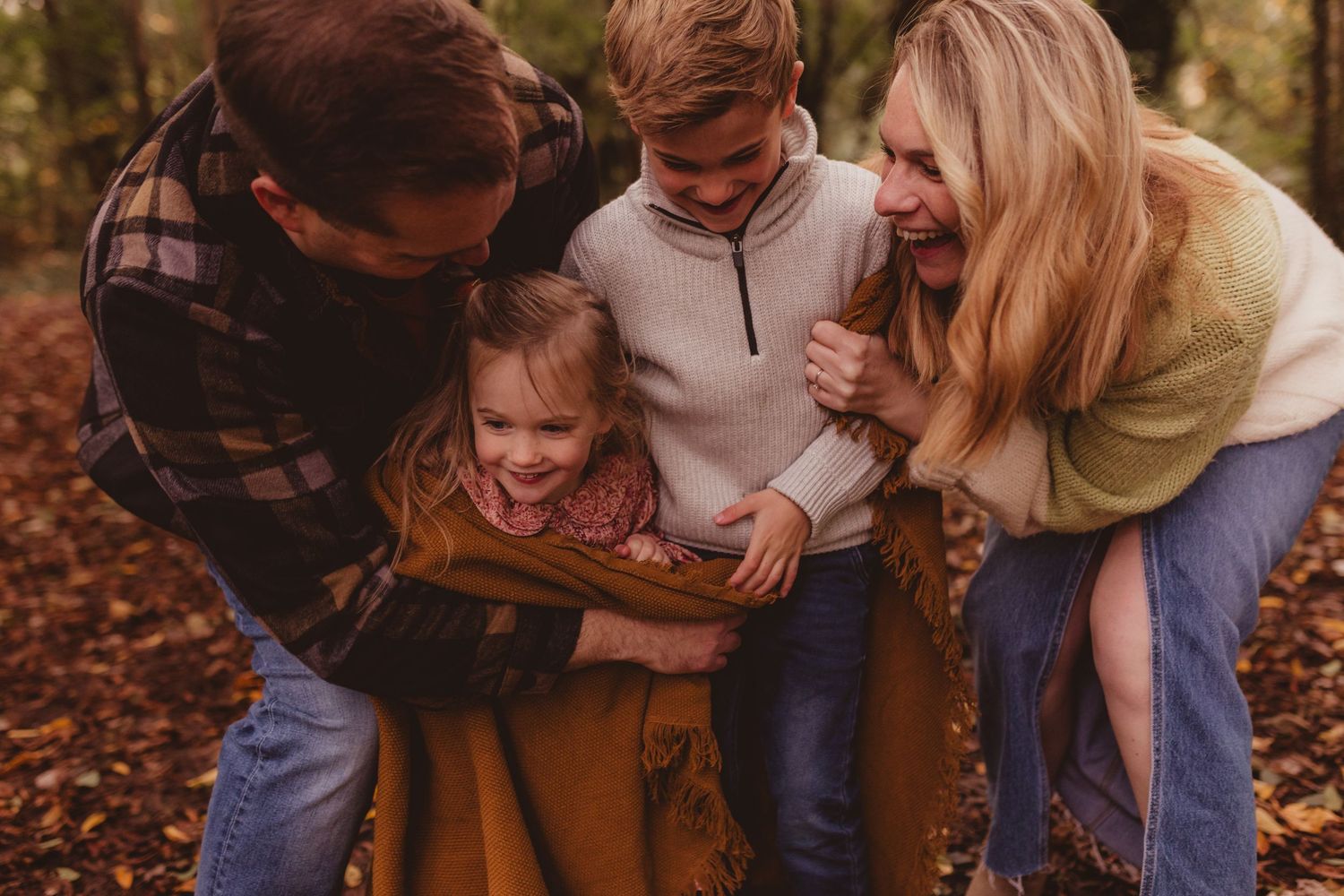 Family of four laughing together outdoors in autumn woods, wrapped in a golden blanket among fallen leaves.