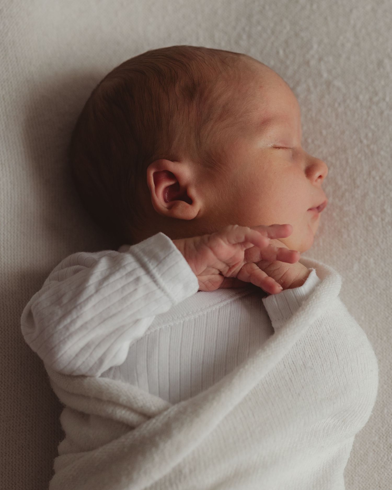 Peaceful newborn baby sleeping swaddled in white blanket, eyes closed, tiny hands folded near chin.