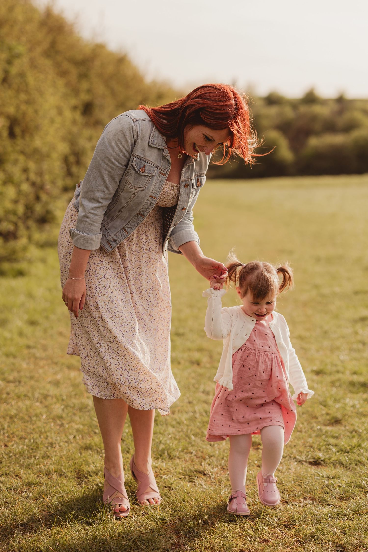 Red-haired mother in floral dress holding toddler girl's hand while walking in a sunny green field.