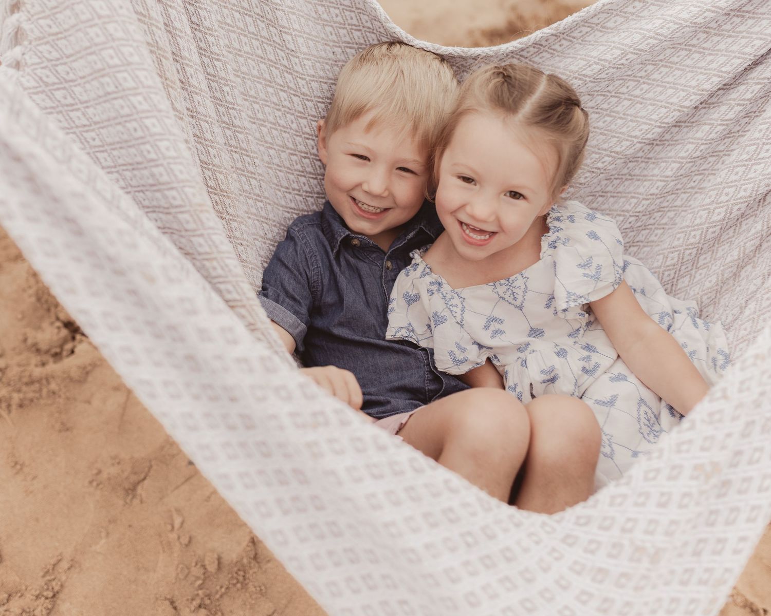 Two smiling children sitting together in a hammock on a sandy beach, wearing casual summer outfits.