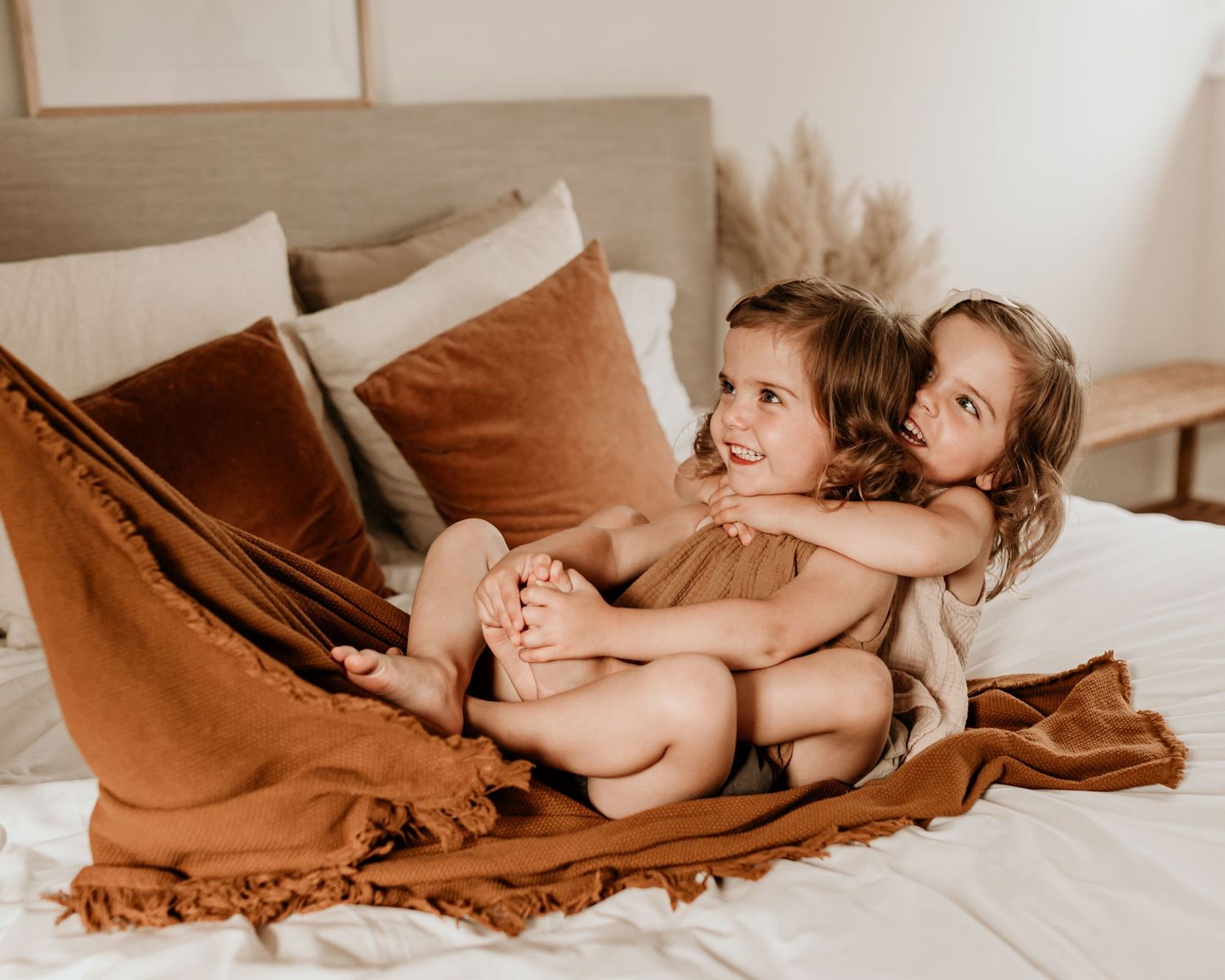 Two young children cuddling on a white bed with rust-colored blanket and pillows in a cozy bedroom.