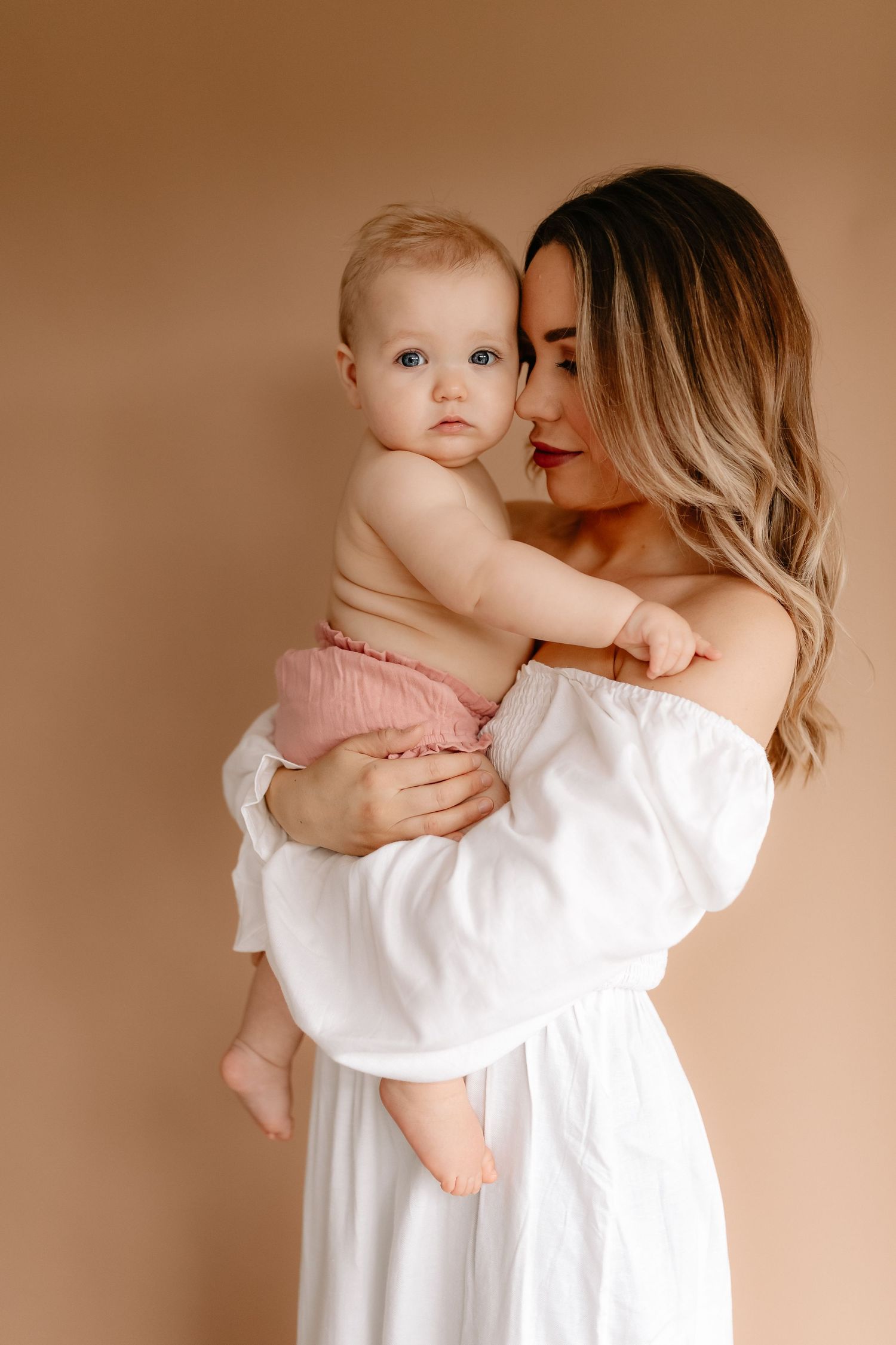 A woman in a white off-shoulder dress tenderly holds her baby girl in pink bloomers against a beige background.