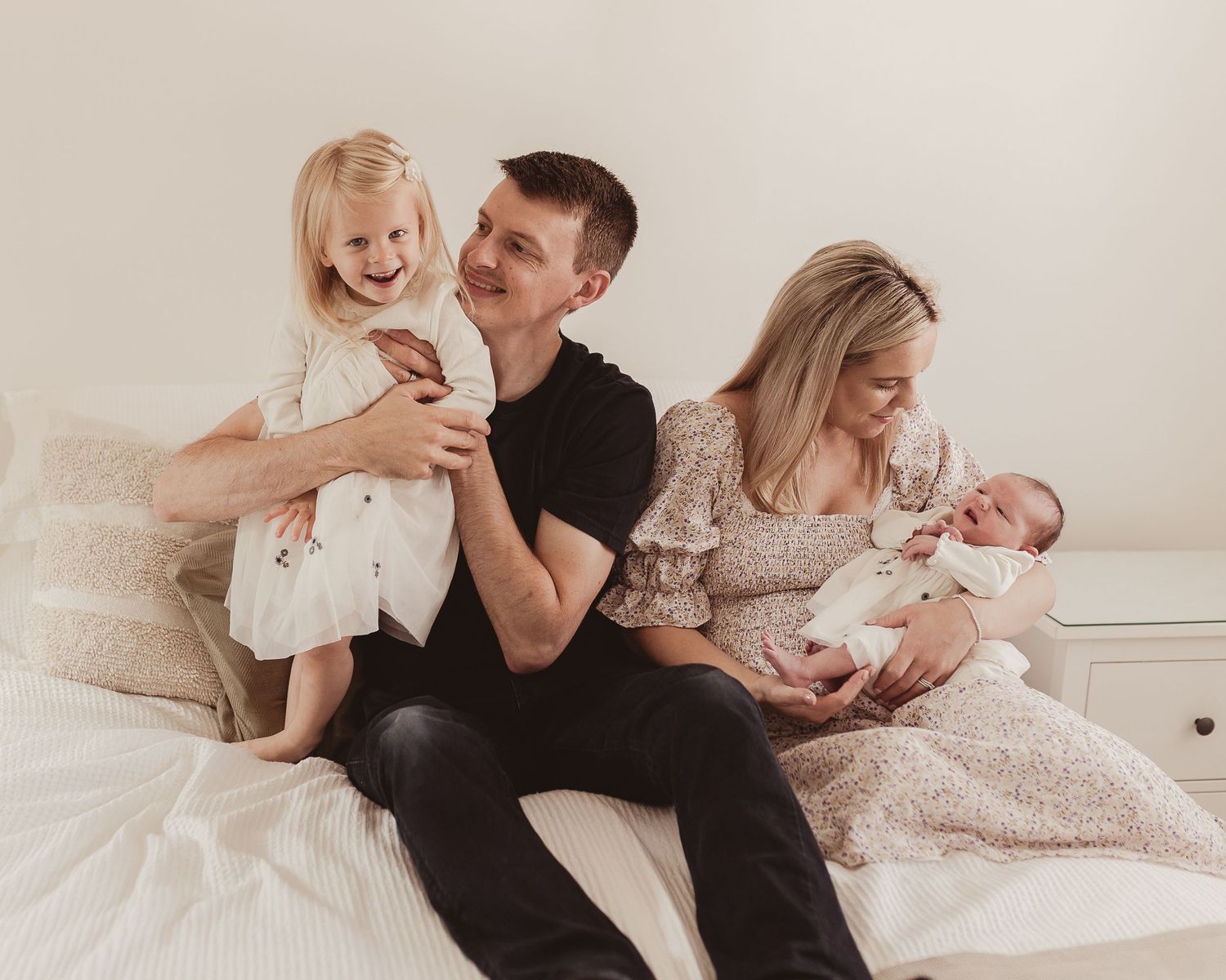Happy family of four sitting on bed, father holding laughing girl while mother cuddles newborn baby.