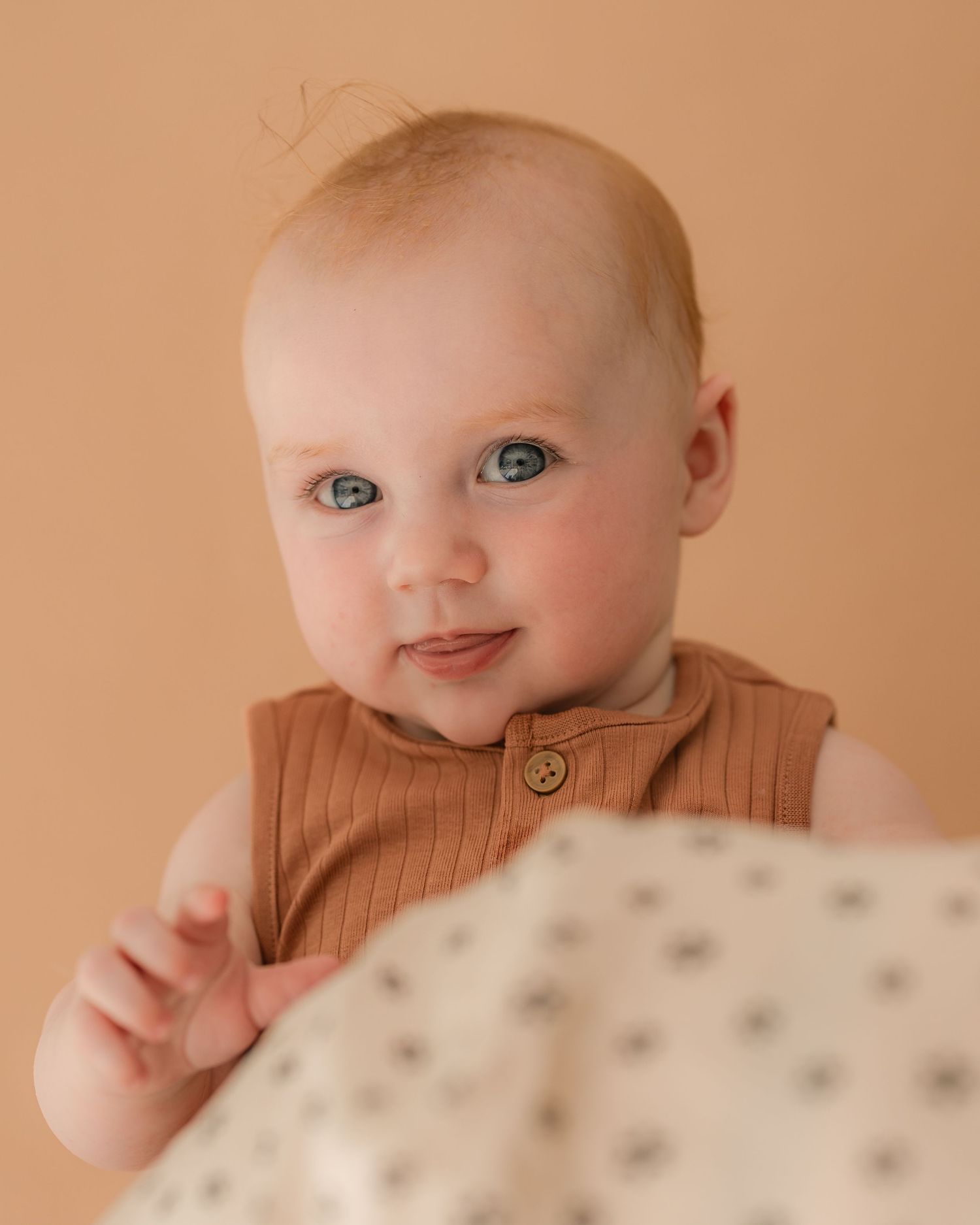 Adorable blue-eyed baby in a brown ribbed romper smiling against a warm beige background.