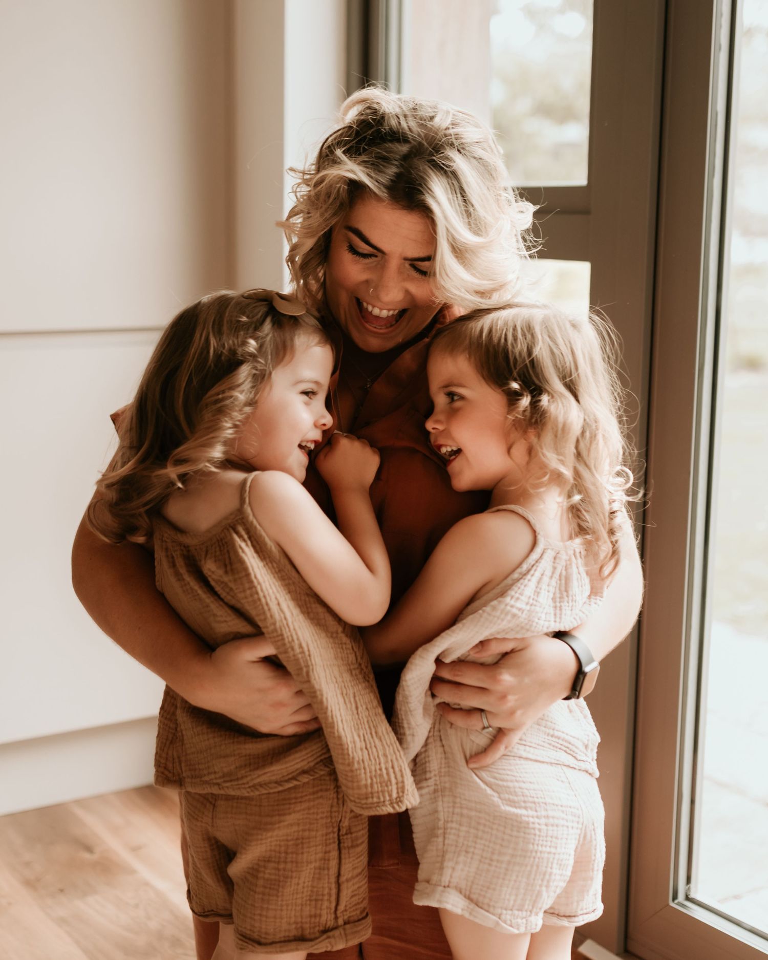 A laughing blonde mother embraces her two young daughters in matching earth-tone outfits near a bright window.