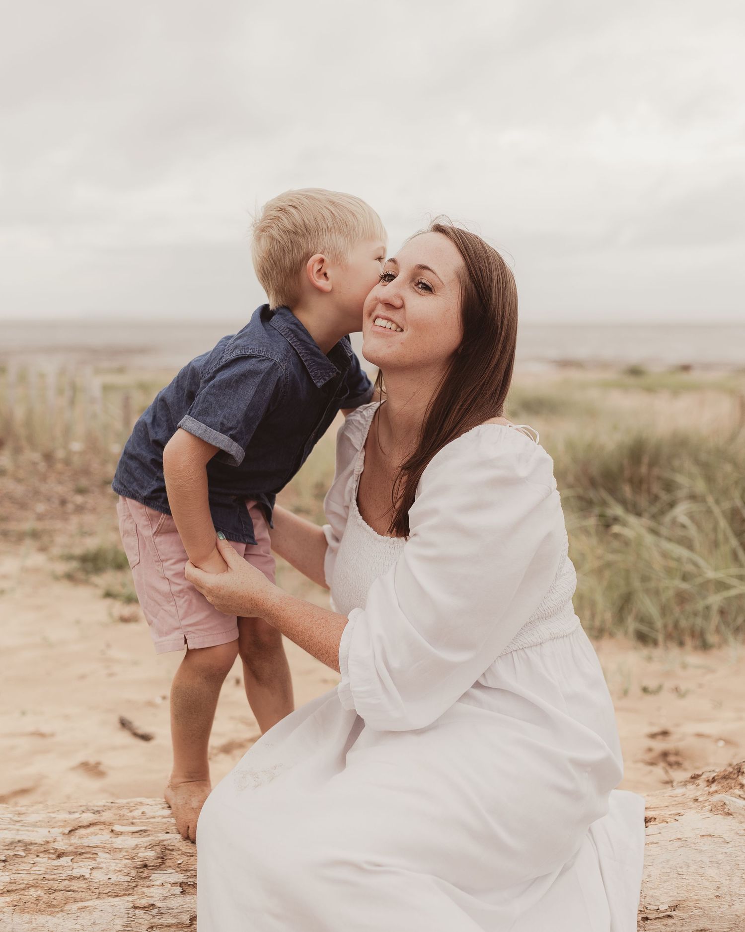 Young boy giving his smiling mother a kiss on the cheek while she crouches on a sandy beach.