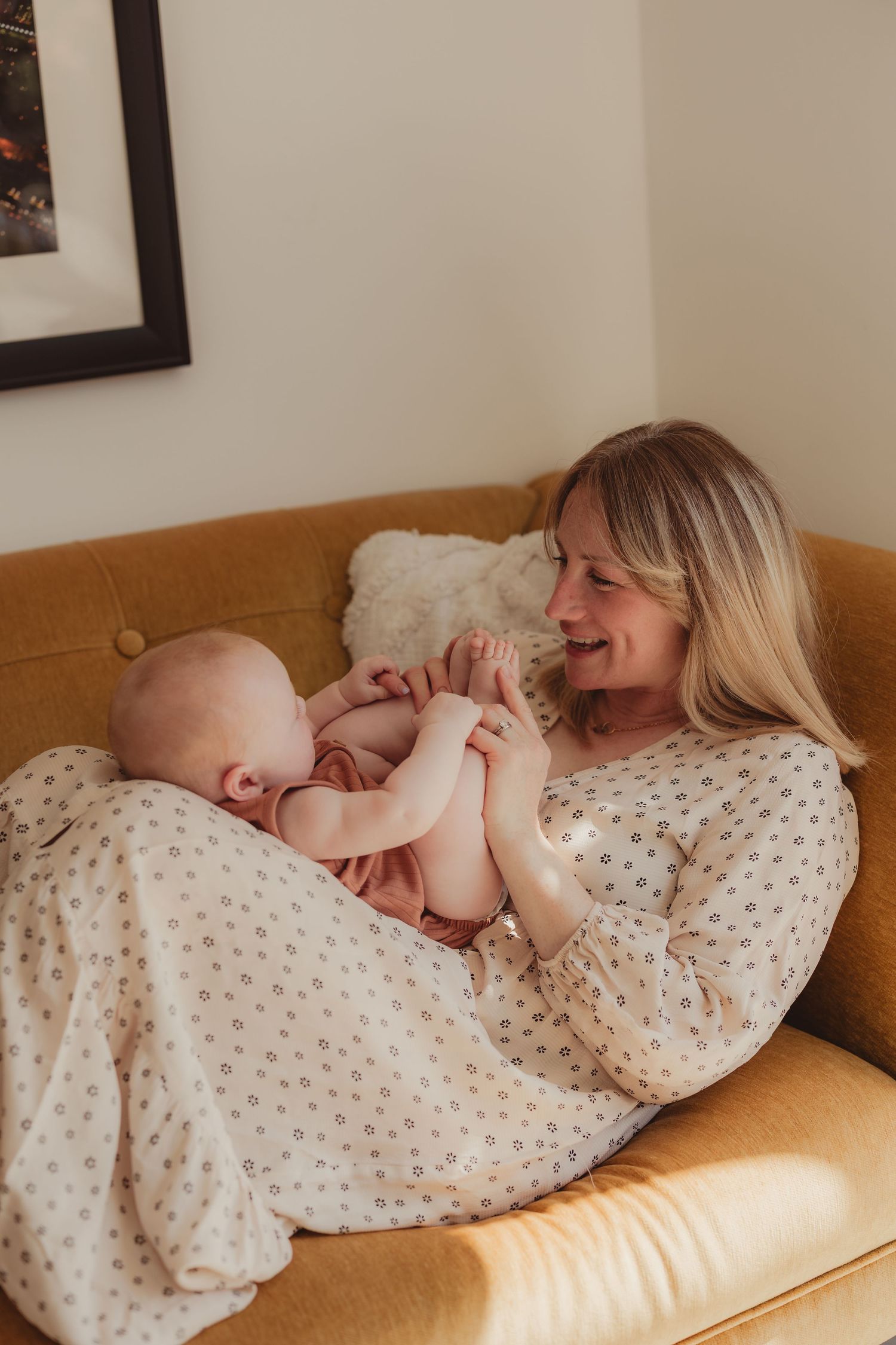 A smiling mother holds her baby on a mustard yellow sofa, playfully interacting in a cozy, warmly lit living room.