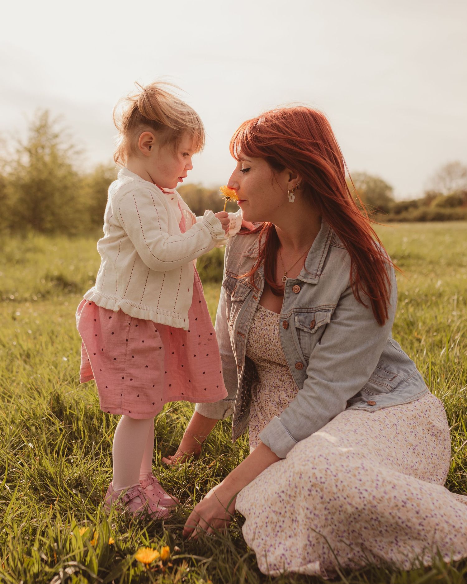 A red-haired mother kneels in a meadow as her toddler daughter offers her a yellow flower to smell.