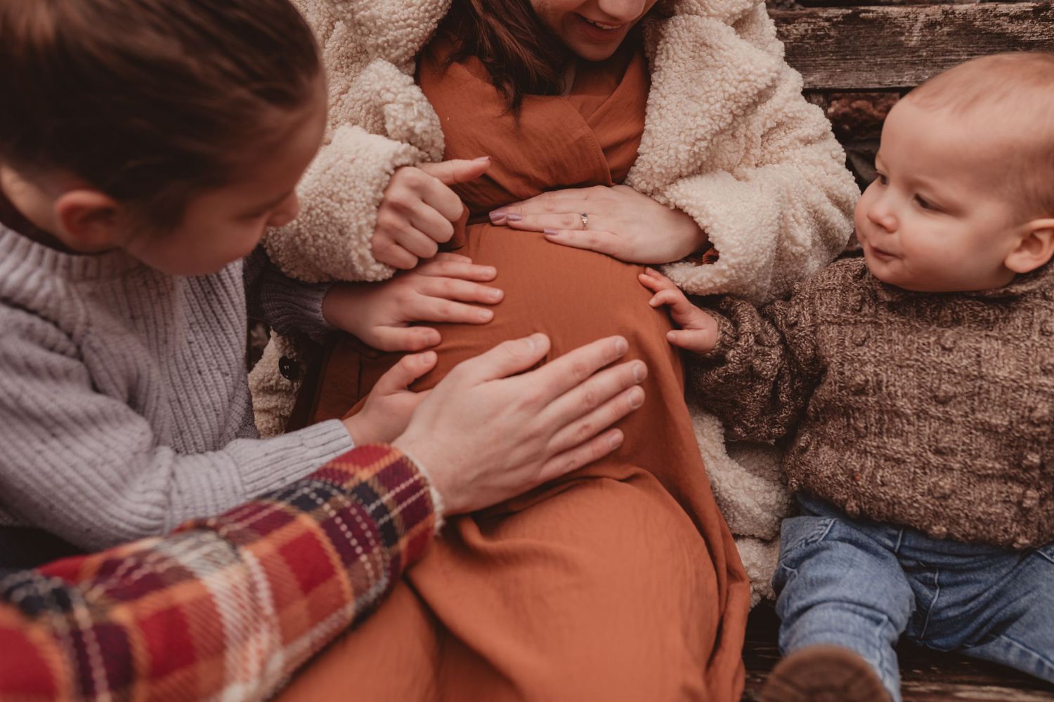 Family touching pregnant woman's belly outdoors in warm autumn tones.