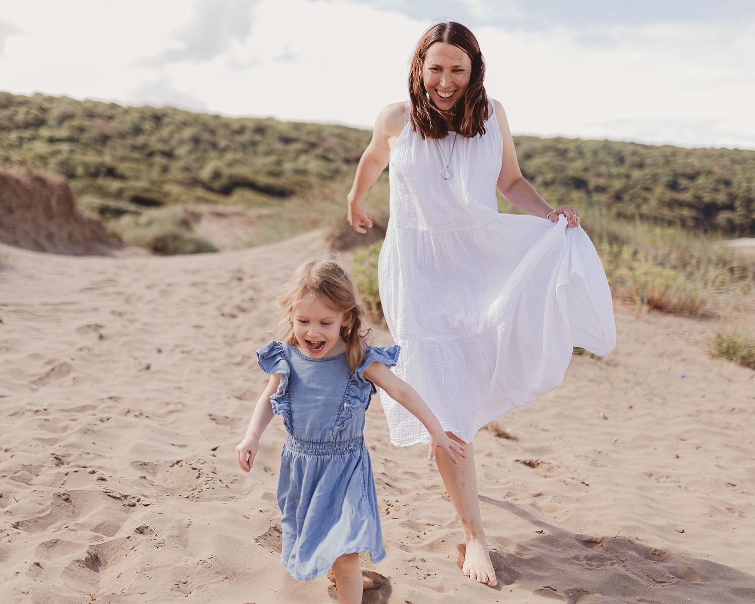 Joyful mother in white dress running with young daughter on sandy beach dunes on cloudy day.