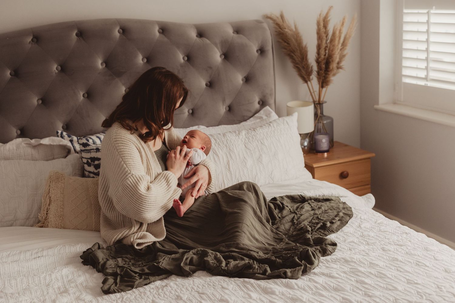 A mother sits on a bed cradling her newborn baby, wrapped in a cozy cream sweater with a dark olive blanket.