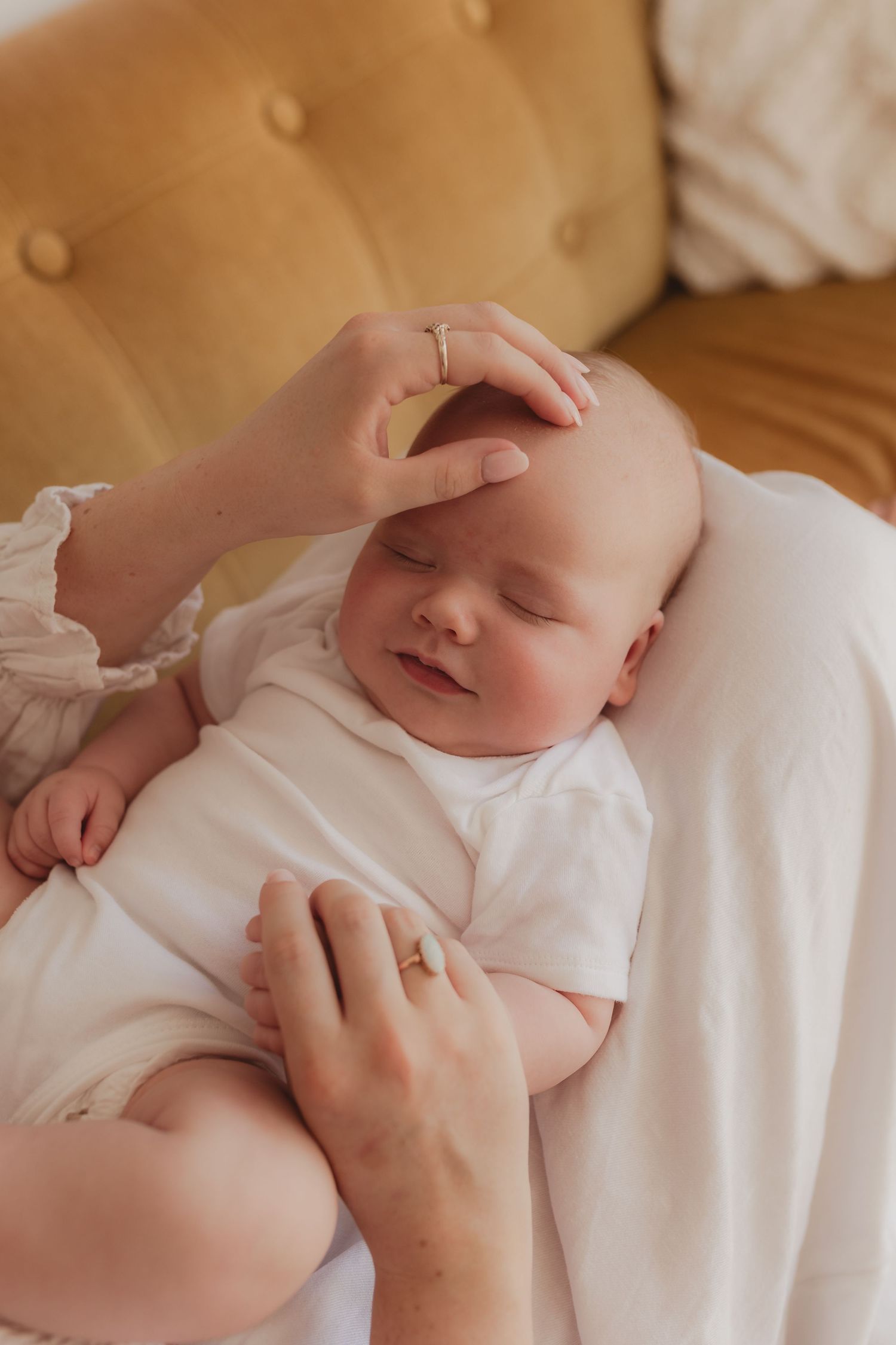A mother gently strokes her sleeping newborn baby dressed in white, cradled in her arms on a golden sofa.