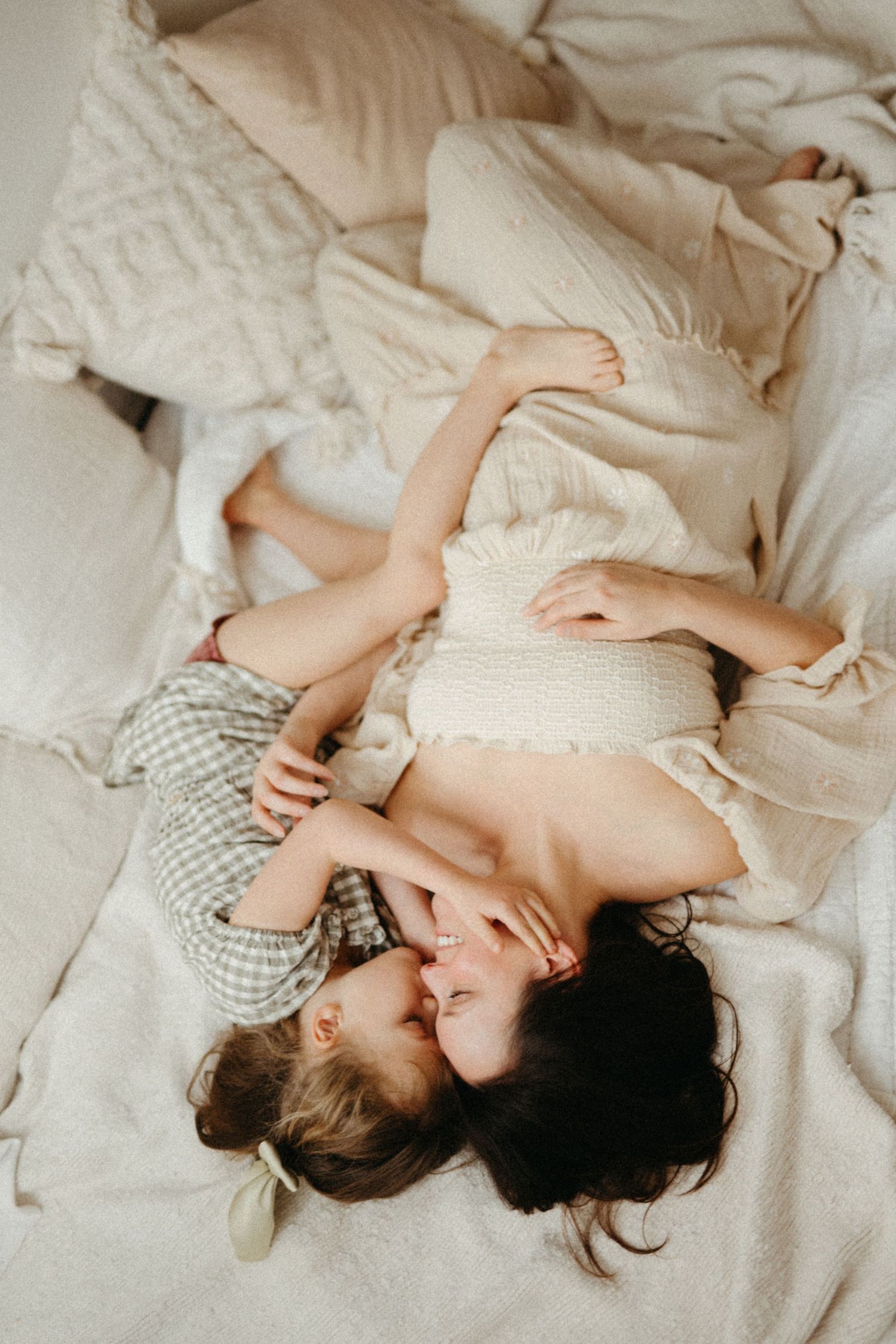 A mother and young daughter share a tender moment lying together on soft beige bedding, smiling face to face.