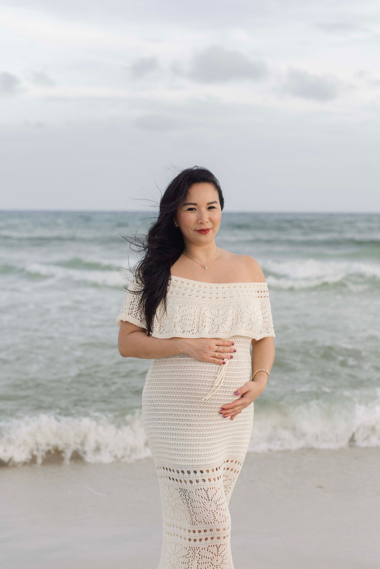 A maternity photoshoot on a cloudy beach with waves crashing in the background, wearing a white crochet dress.