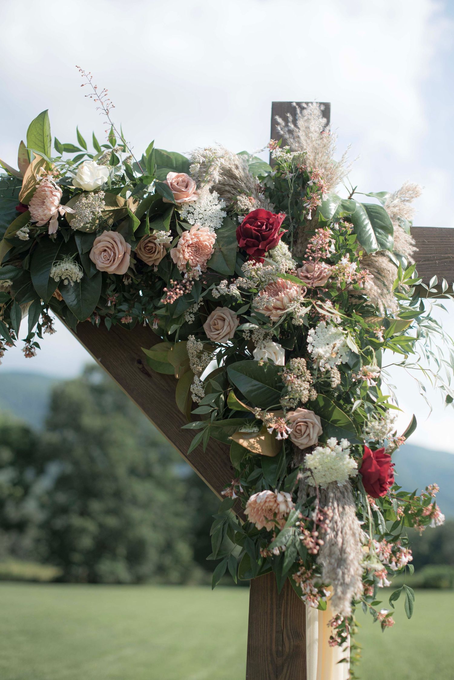 Romantic floral arch arrangement with pink roses, white blooms and greenery against a pastoral landscape.