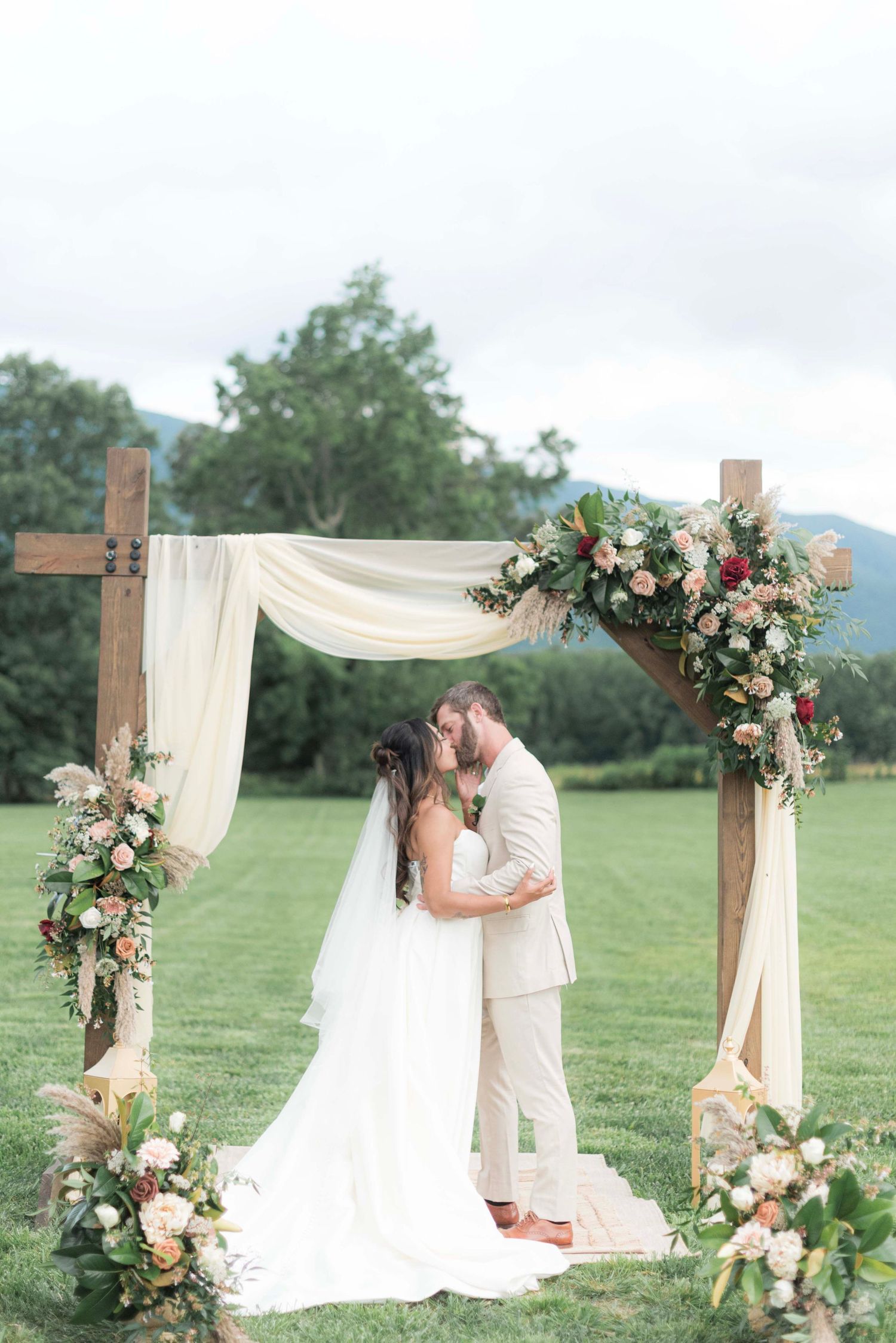 A romantic outdoor wedding ceremony with a wooden arch draped in white fabric and decorated with pink and white flowers.