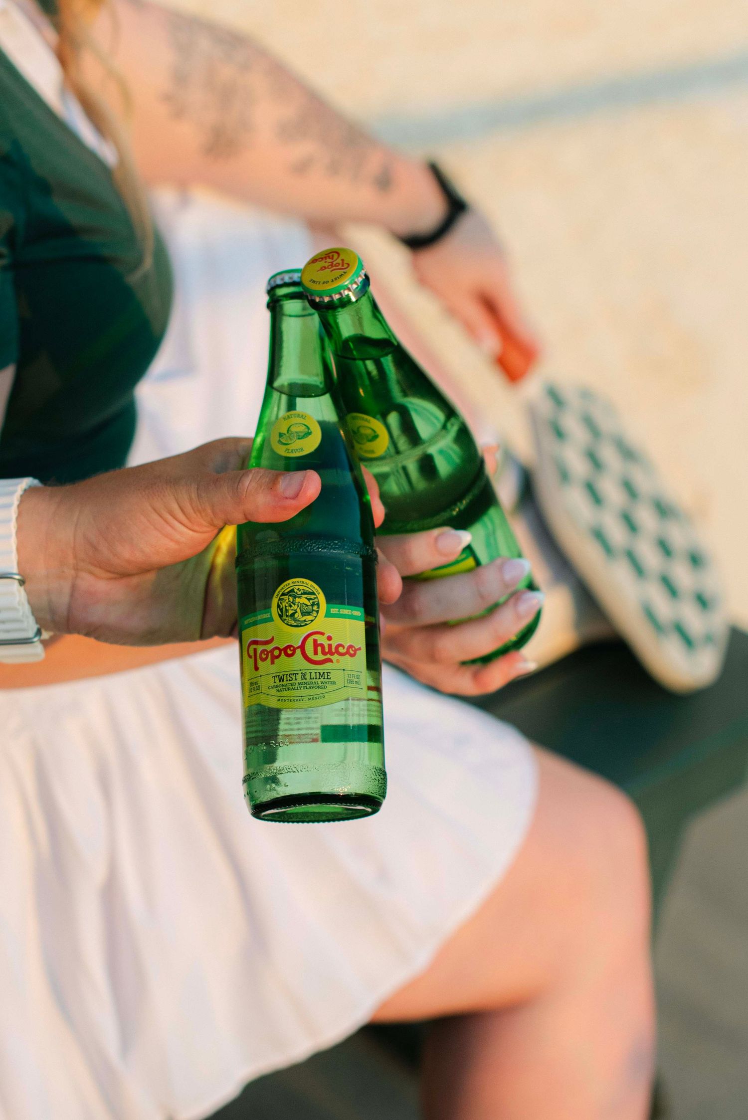 Close-up shot of hands holding green glass Topo Chico bottles against a light background.