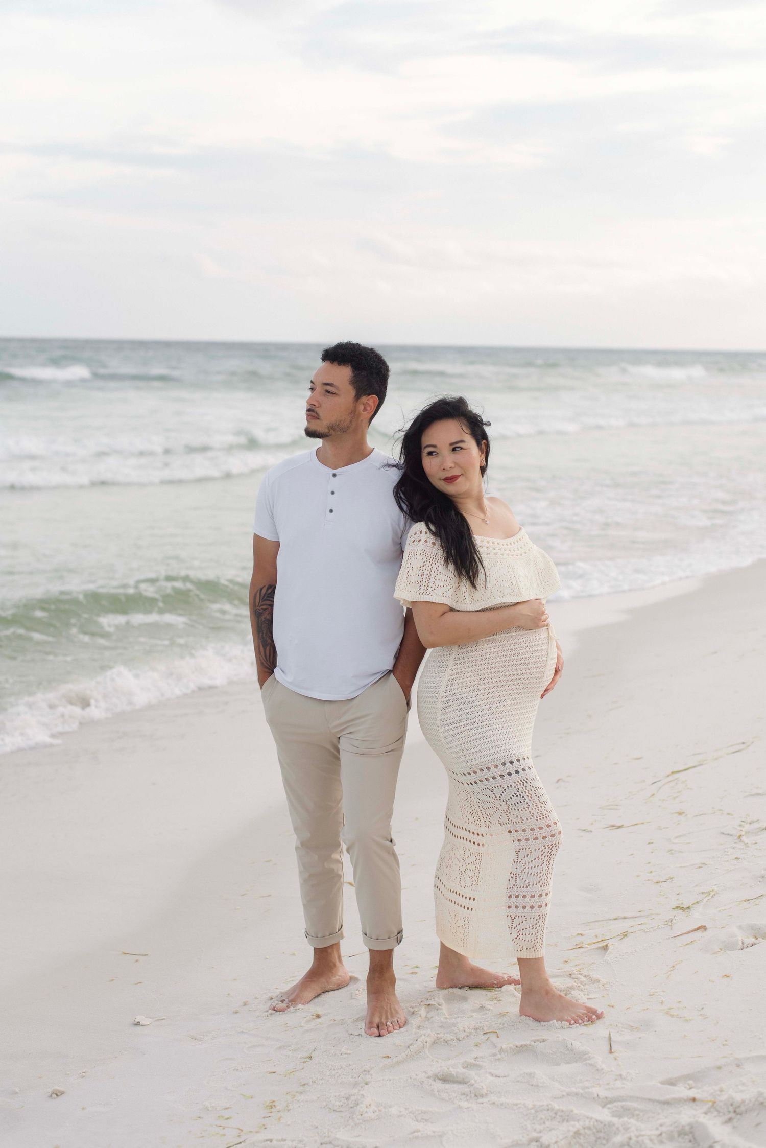 A couple in coordinated white and beige outfits walking along a peaceful beach shoreline during sunset.