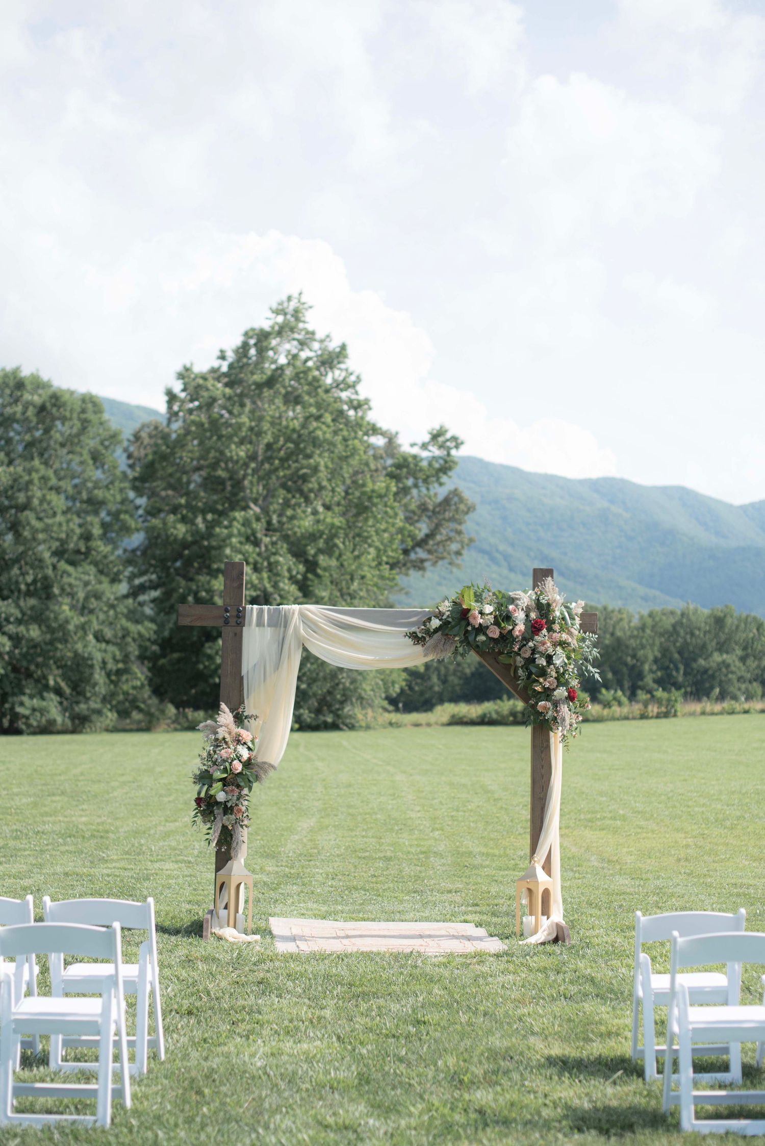 Rustic outdoor wedding ceremony setup with white chairs and floral arch draped in sheer fabric against mountain backdrop.