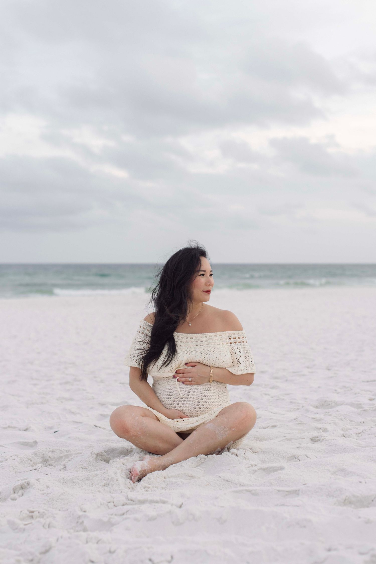 Woman in white off-shoulder dress poses gracefully on pristine white sandy beach with ocean waves in background.