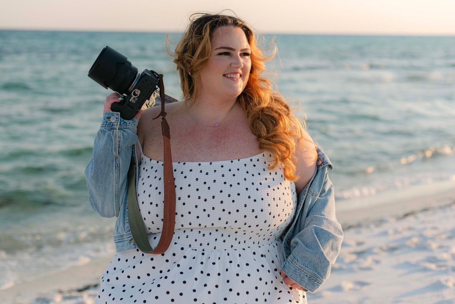 Photographer with multiple cameras in denim jacket at beach during golden hour.