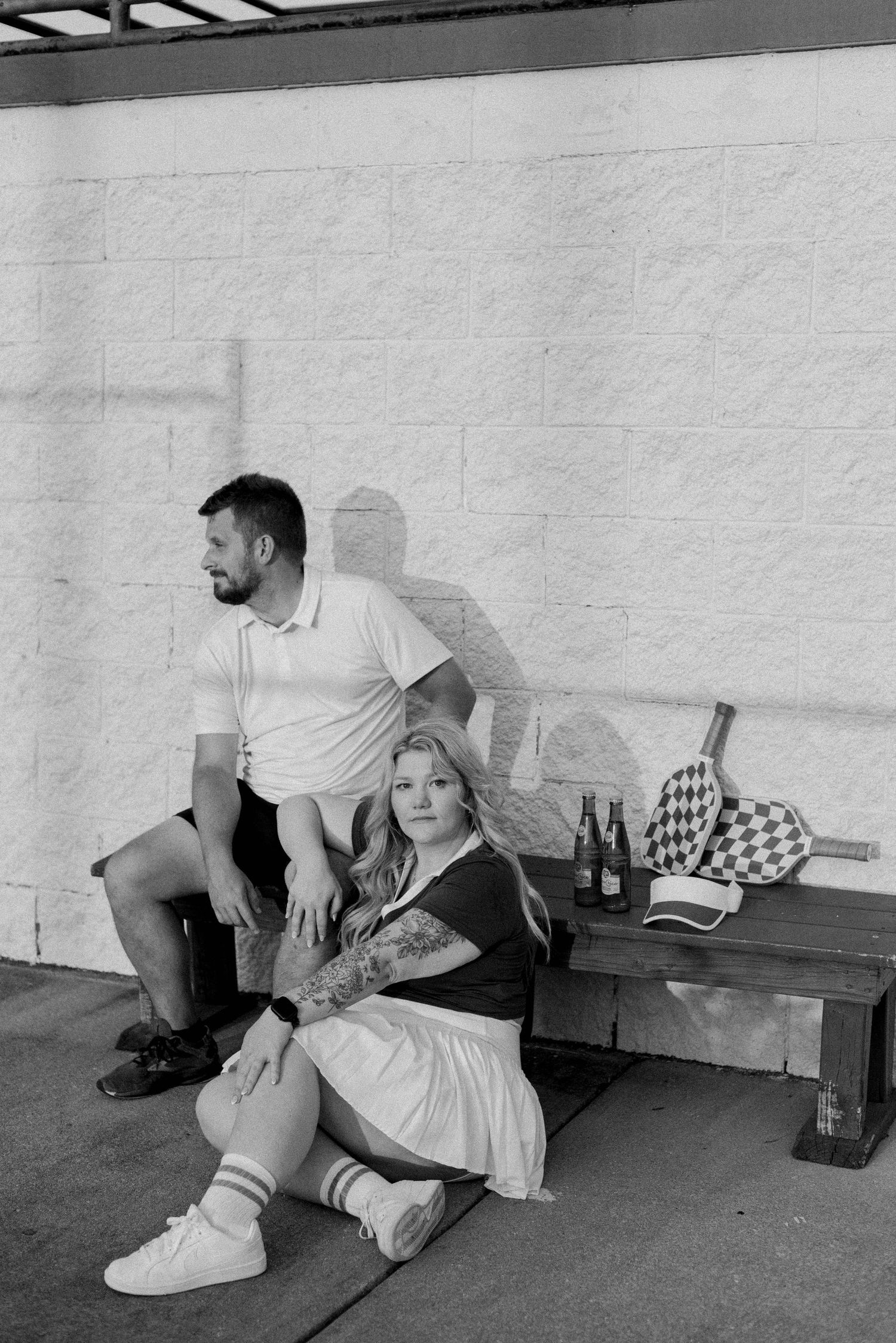 Black and white photo of people relaxing on a bench with bottles and a checkered bag nearby.