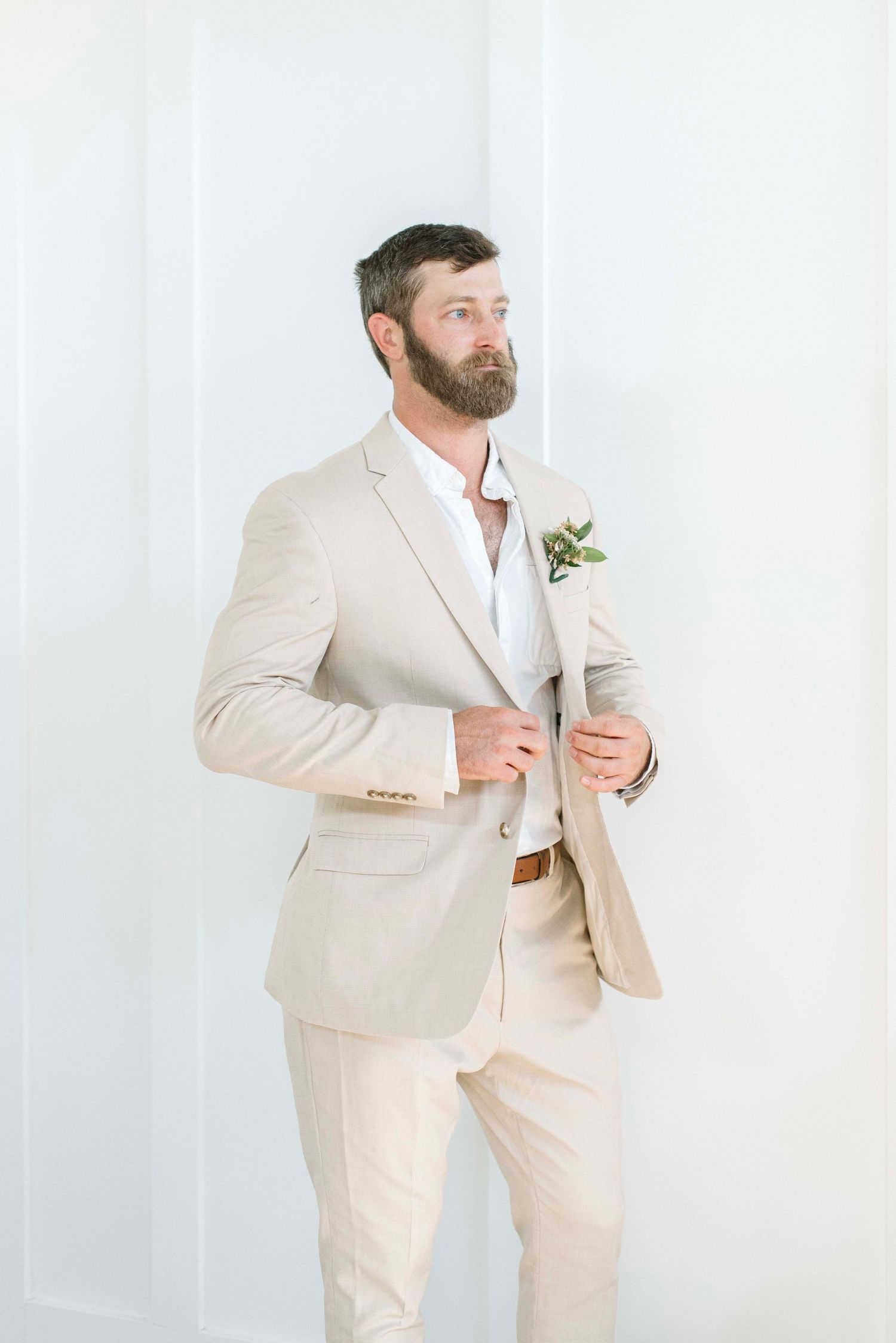 A groom in a cream linen suit and boutonniere poses against a white backdrop in a series of candid shots.
