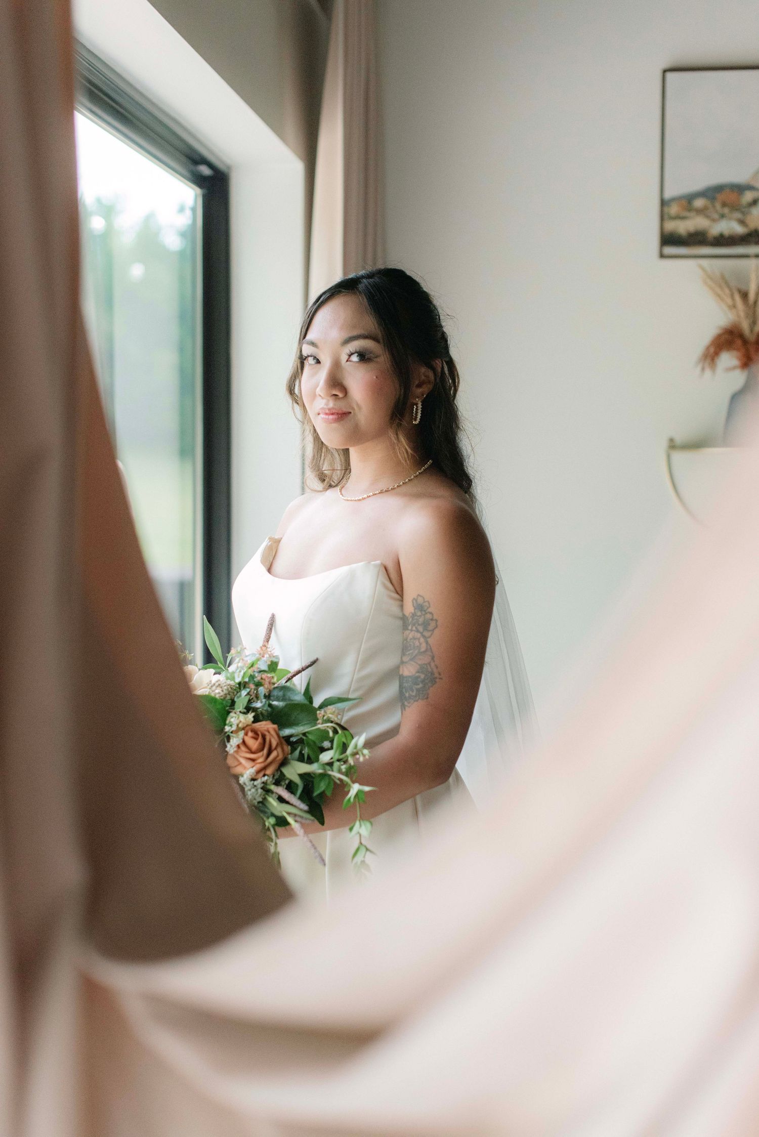 A bride in a white dress stands by a window holding a floral bouquet during her wedding day preparations.