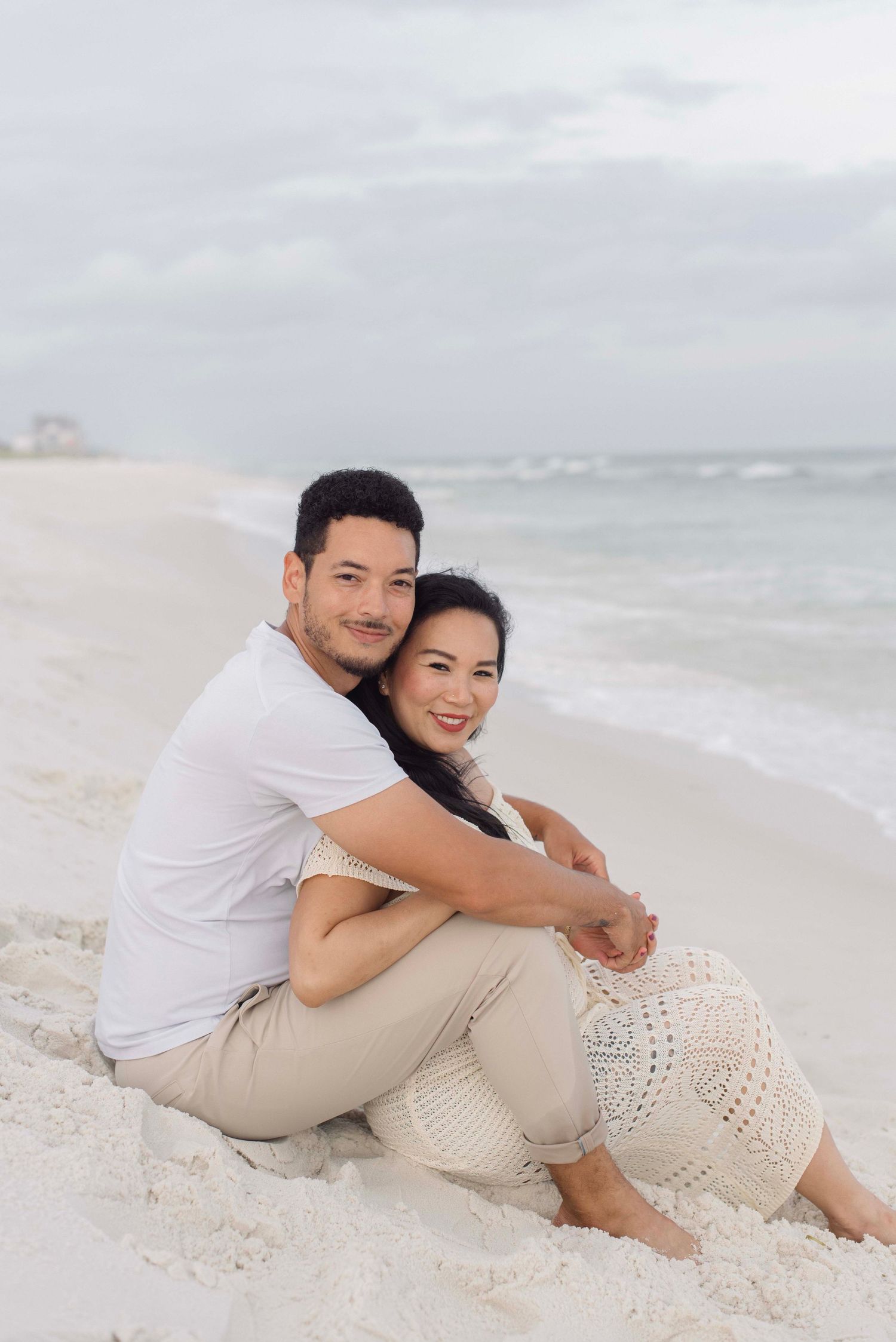 A couple embraces while sitting together on a white sandy beach with ocean waves in the background.