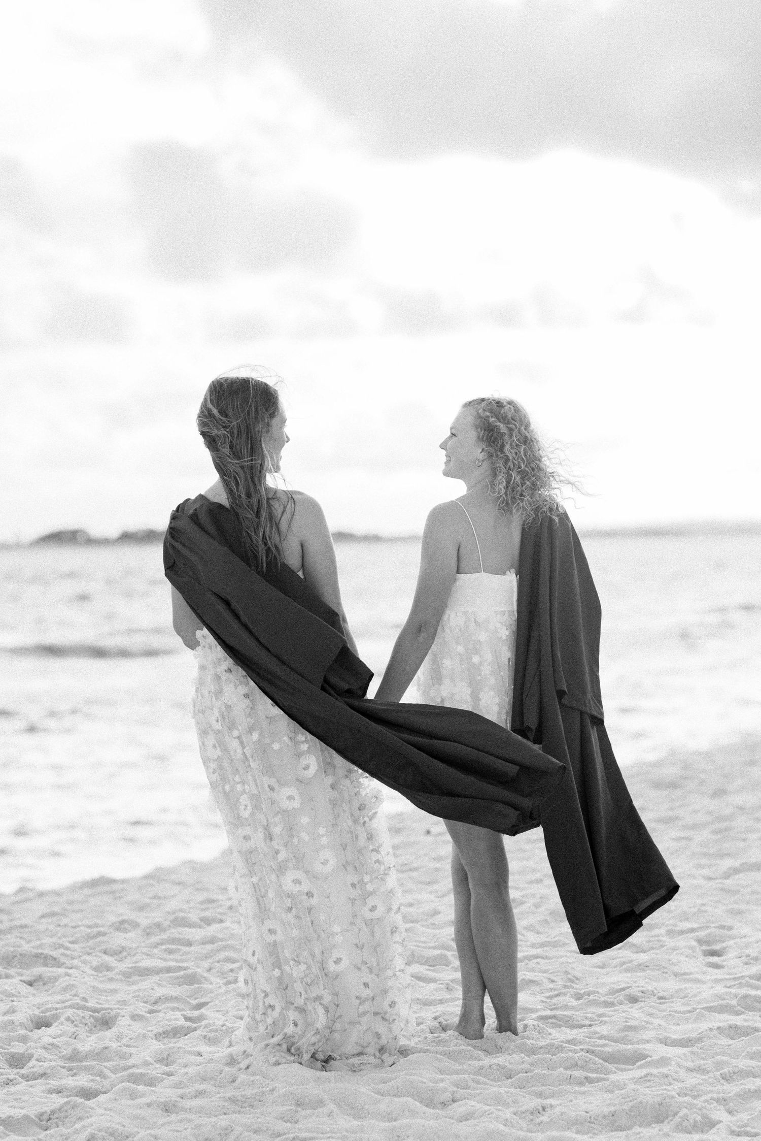 A couple stands together on a sandy beach at sunset in black and white.
