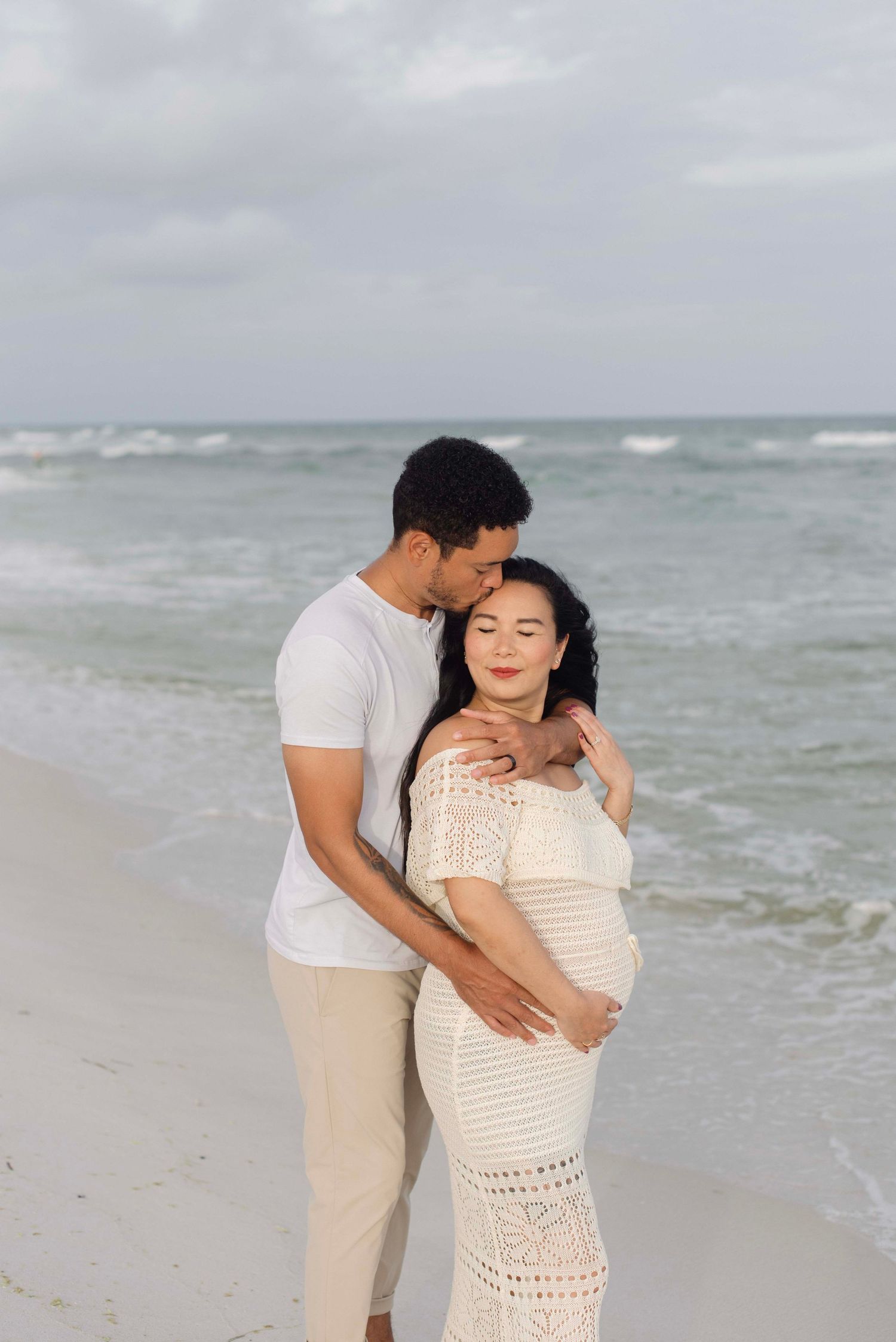 Romantic maternity photo of couple embracing on cloudy beach in white and beige outfits.