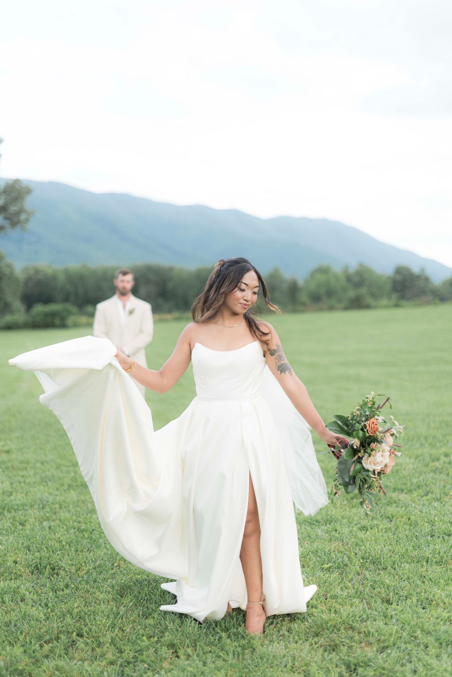 A flowing white wedding dress billows gracefully in the wind as someone walks through a green mountain meadow.