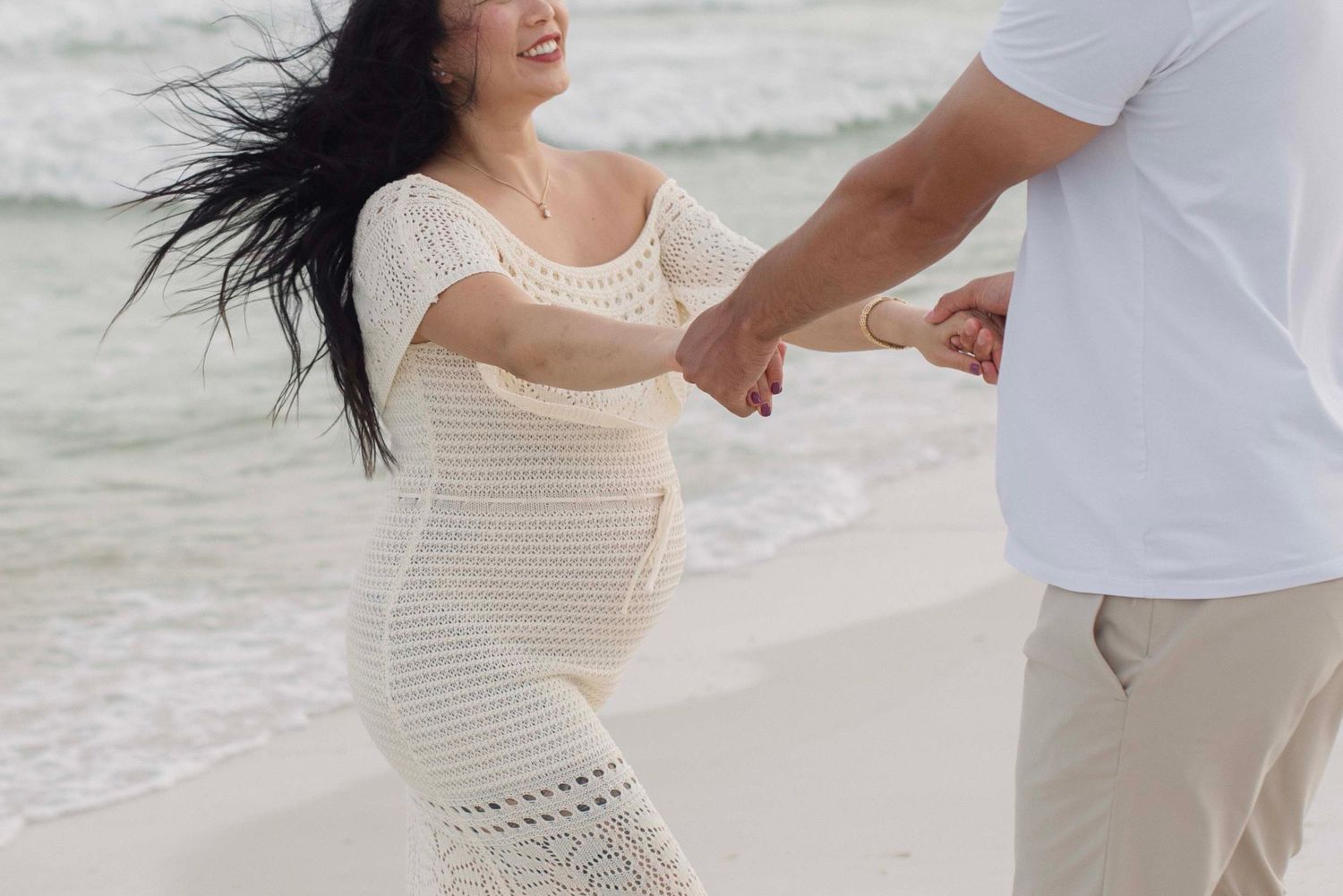 A couple in white outfits dances joyfully together on a beach with waves in the background.