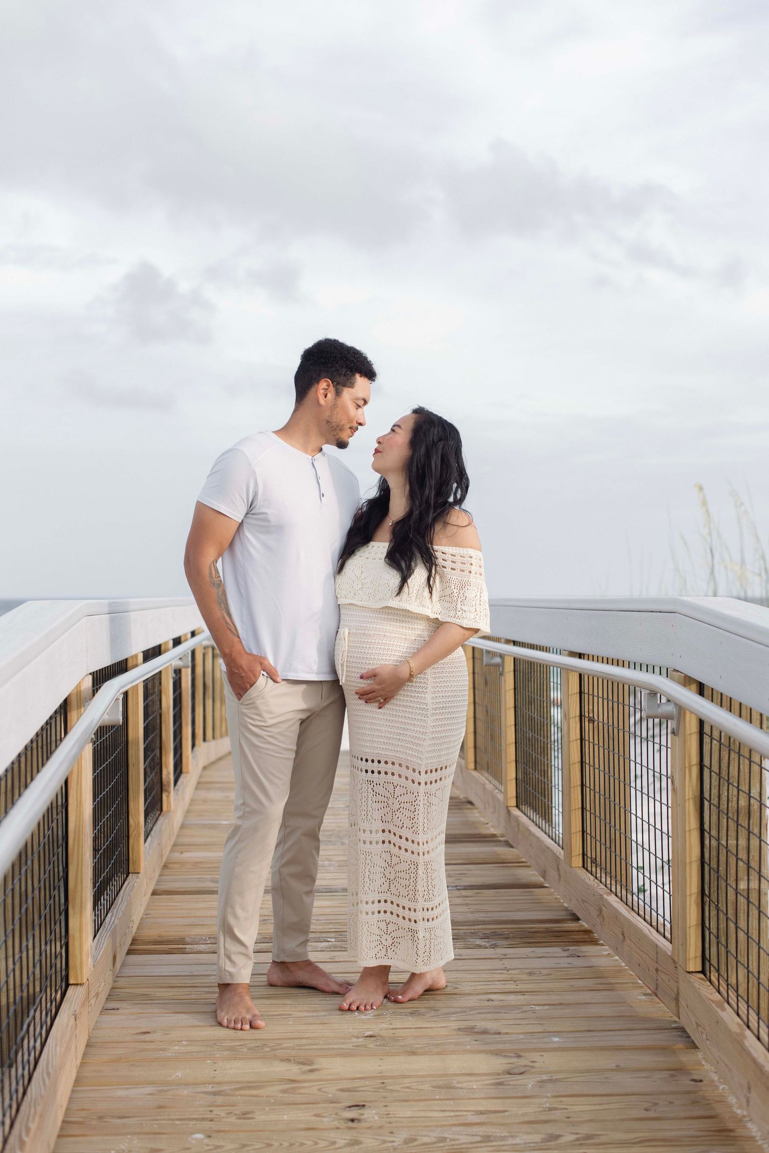 A couple in white outfits stands barefoot on a wooden boardwalk overlooking a cloudy coastal view.