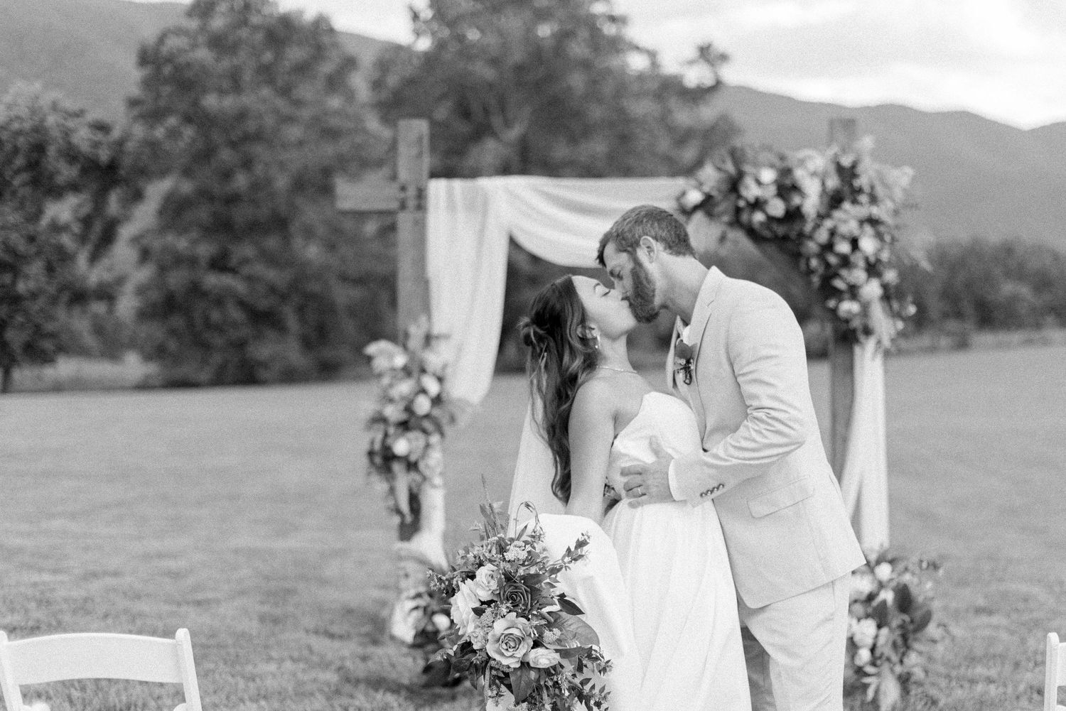 A romantic outdoor wedding ceremony features a white floral arch and elegant seating arrangement in a scenic field.
