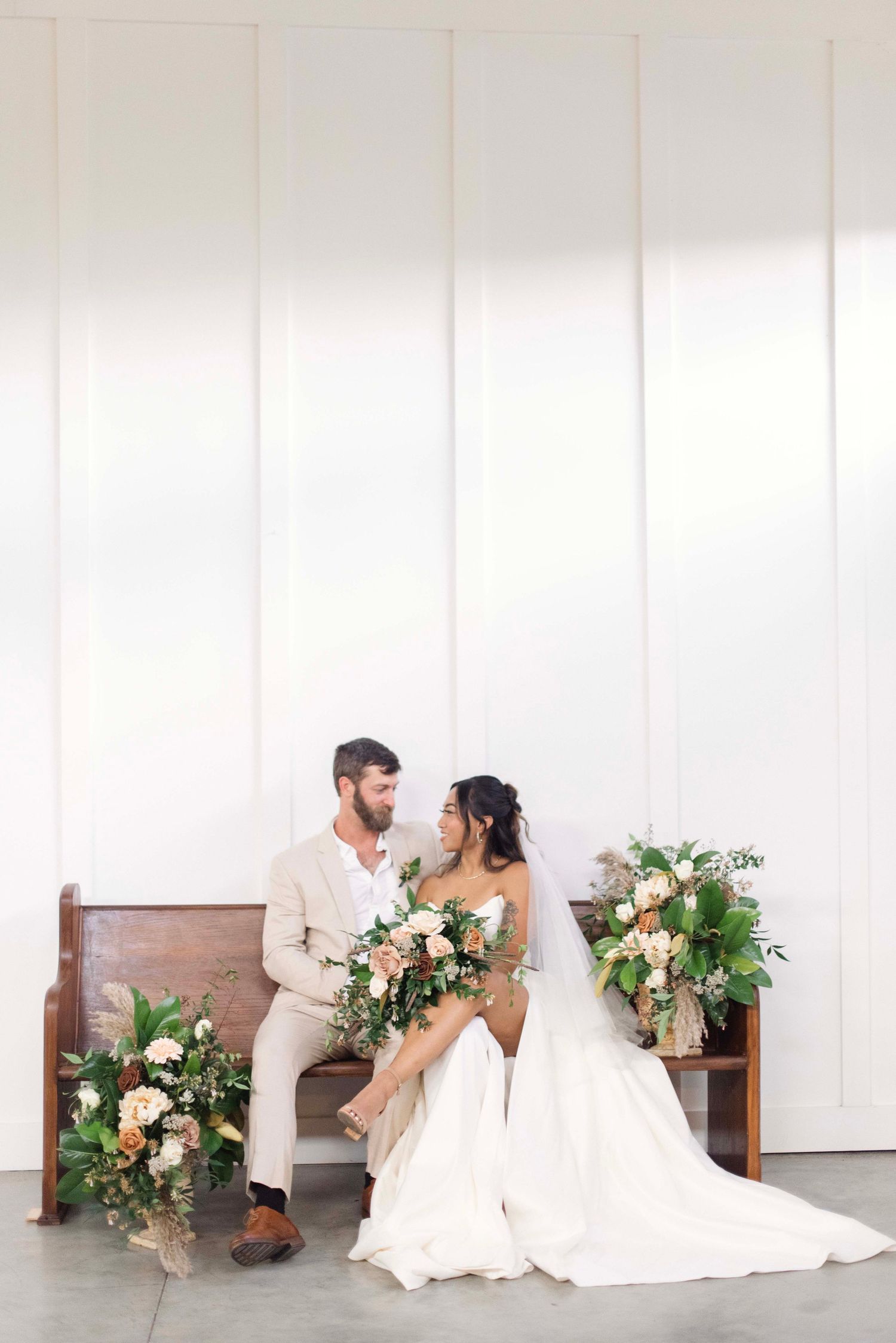 Romantic wedding couple poses intimately on white bench with elegant floral arrangements in bright indoor setting.