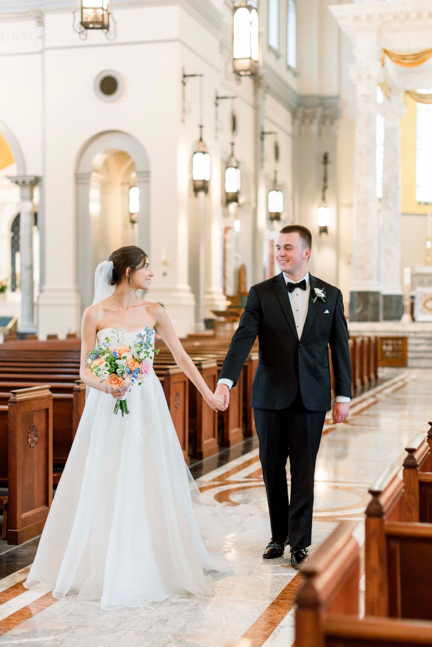 A bride and groom share a tender moment in an elegant white church with wooden pews and hanging lanterns.