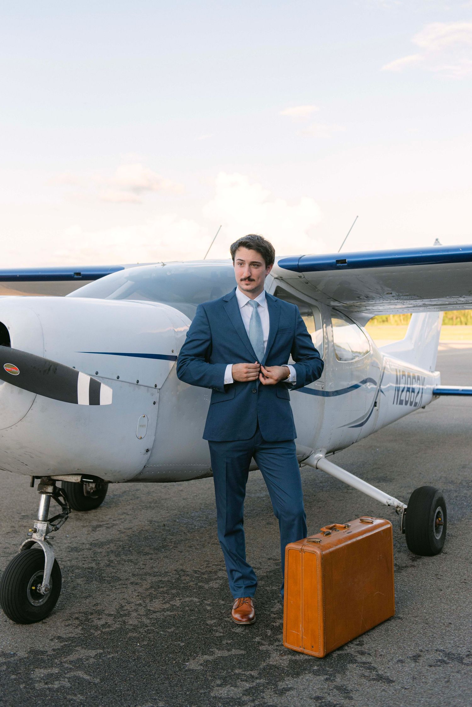 A groom in a blue suit stands next to a small aircraft with vintage brown luggage.