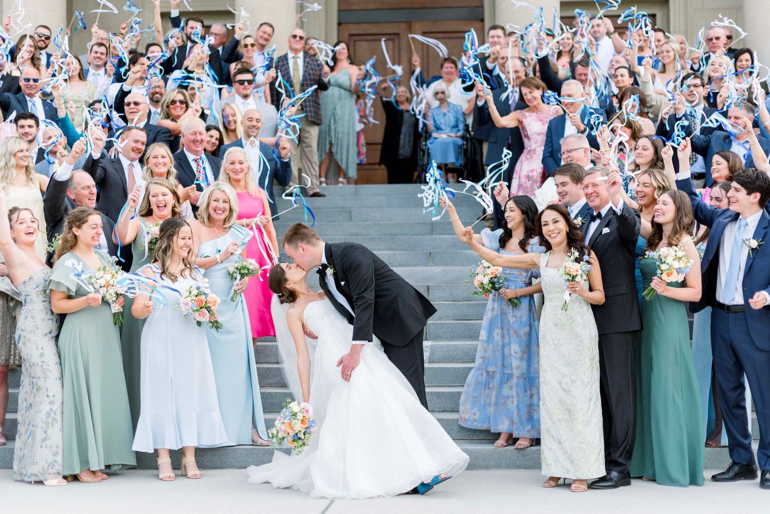 A joyful wedding celebration with guests cheering outside a church as a couple shares a romantic dip kiss on the steps.