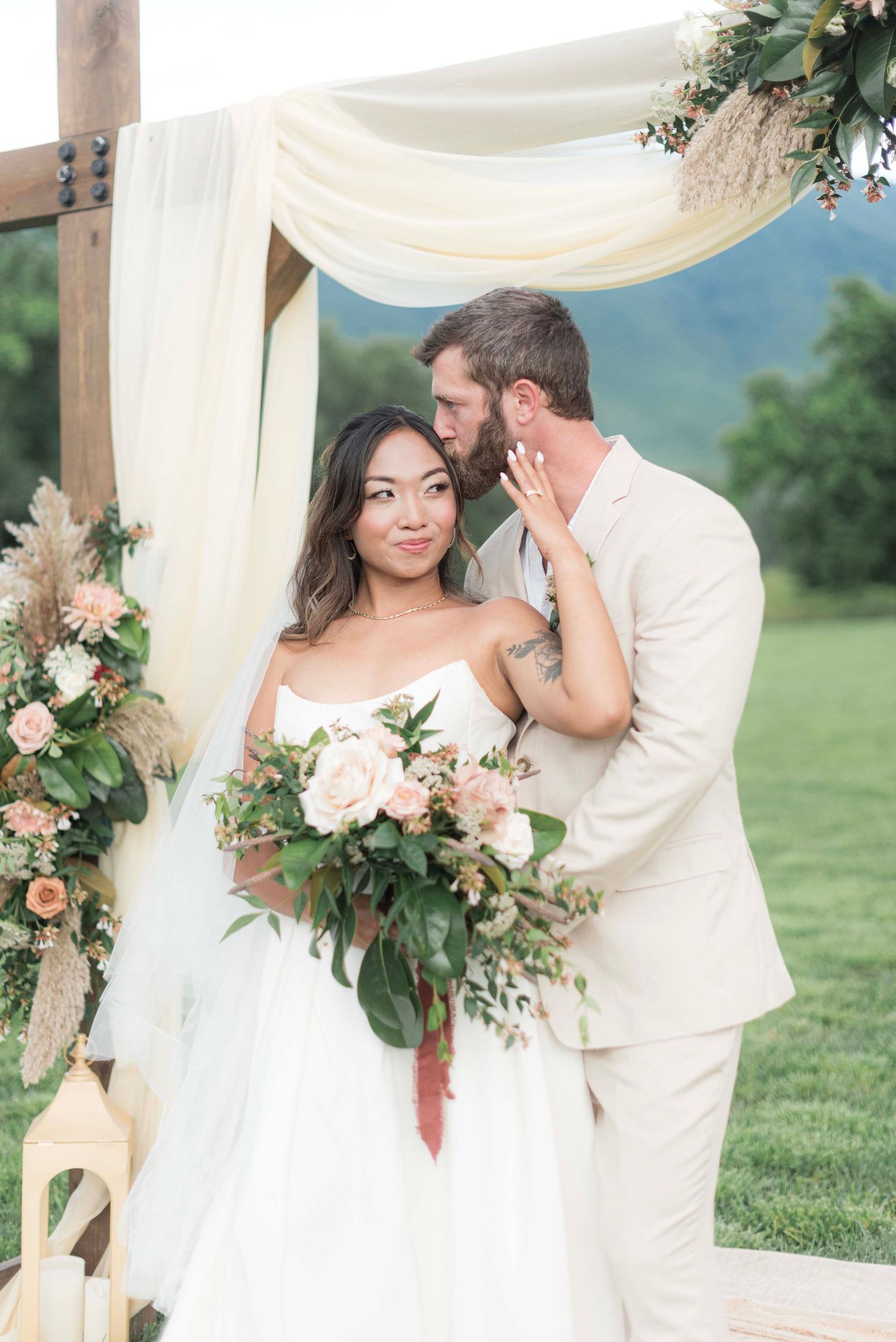 A romantic wedding couple embraces under a draped white canopy decorated with pink flowers and greenery in an outdoor setting.