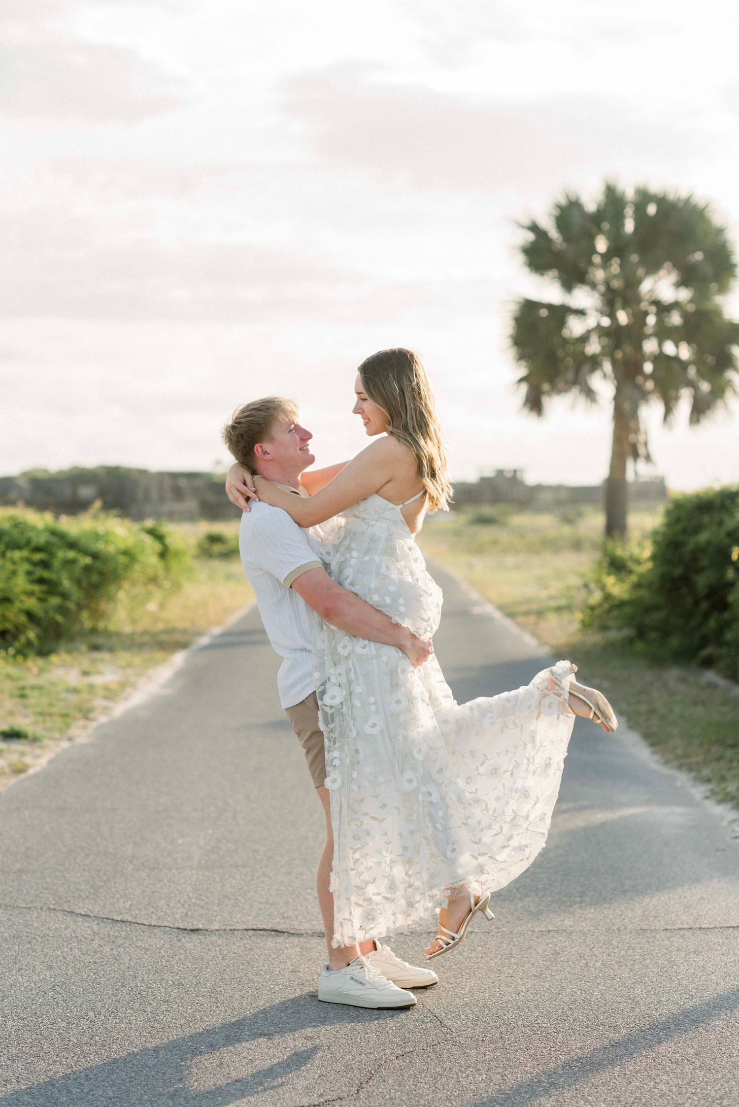 Playful couple in white attire spinning and dancing on a coastal road at sunset.