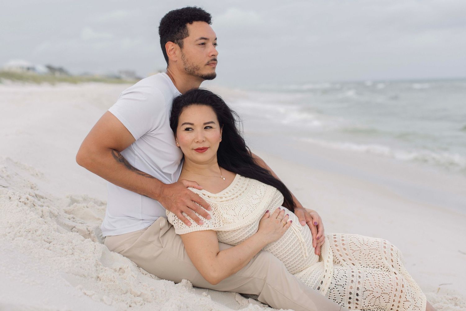 A couple in casual beachwear posing romantically on the sand with the ocean behind them.