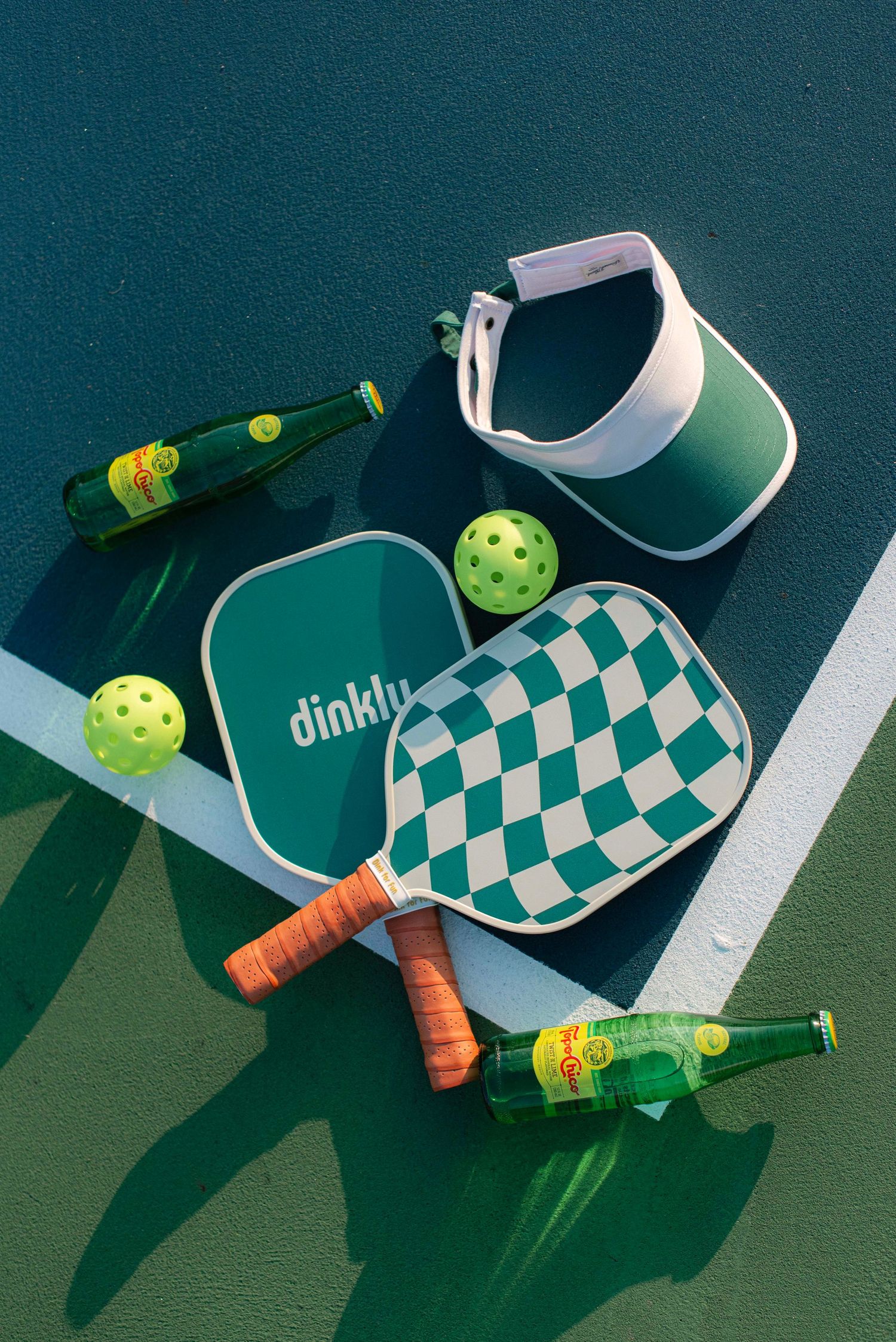Pickleball equipment including paddles, balls, and beverages arranged in a creative flat lay composition on green surface.
