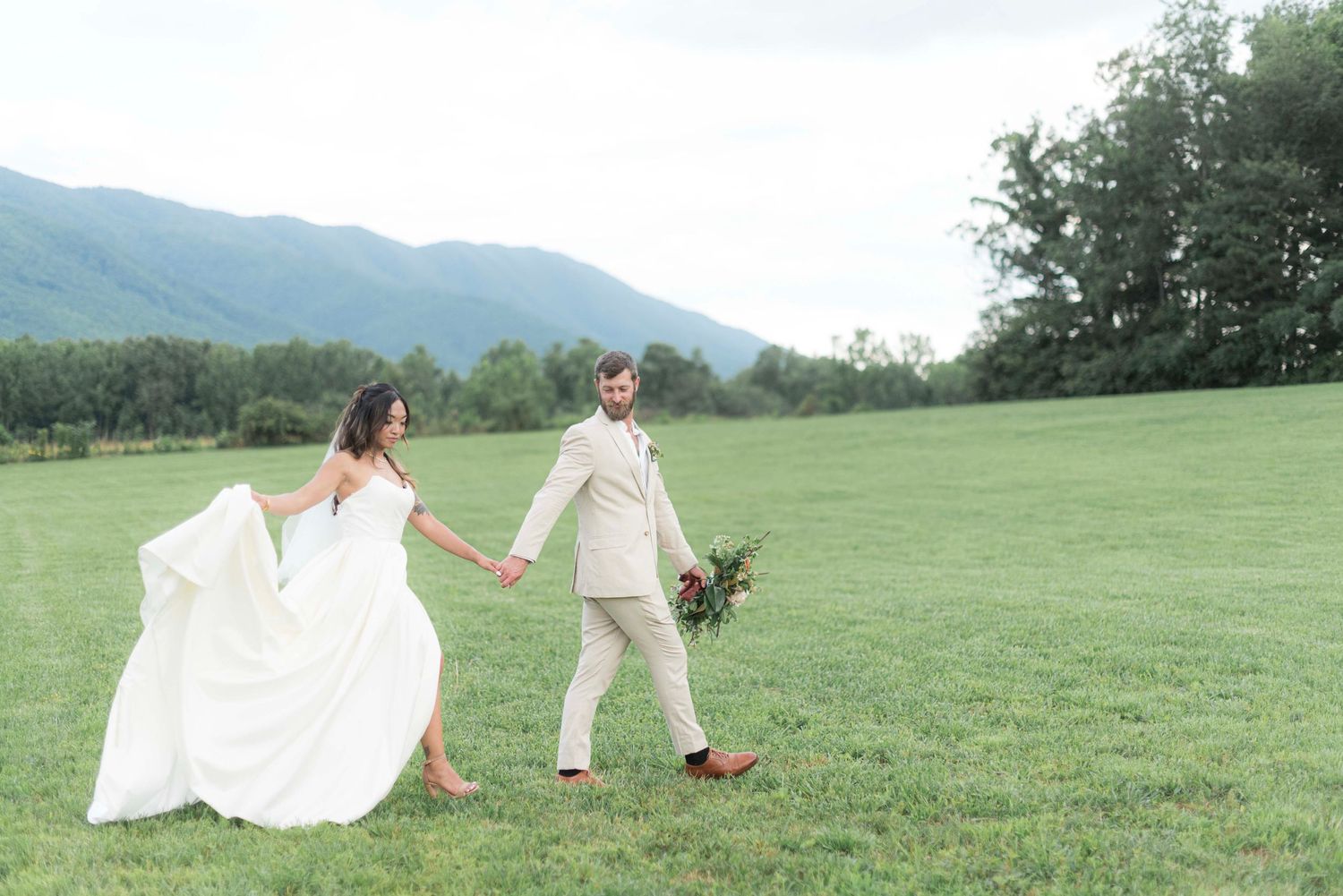 A couple in wedding attire walks through a lush green meadow with mountains in the background.