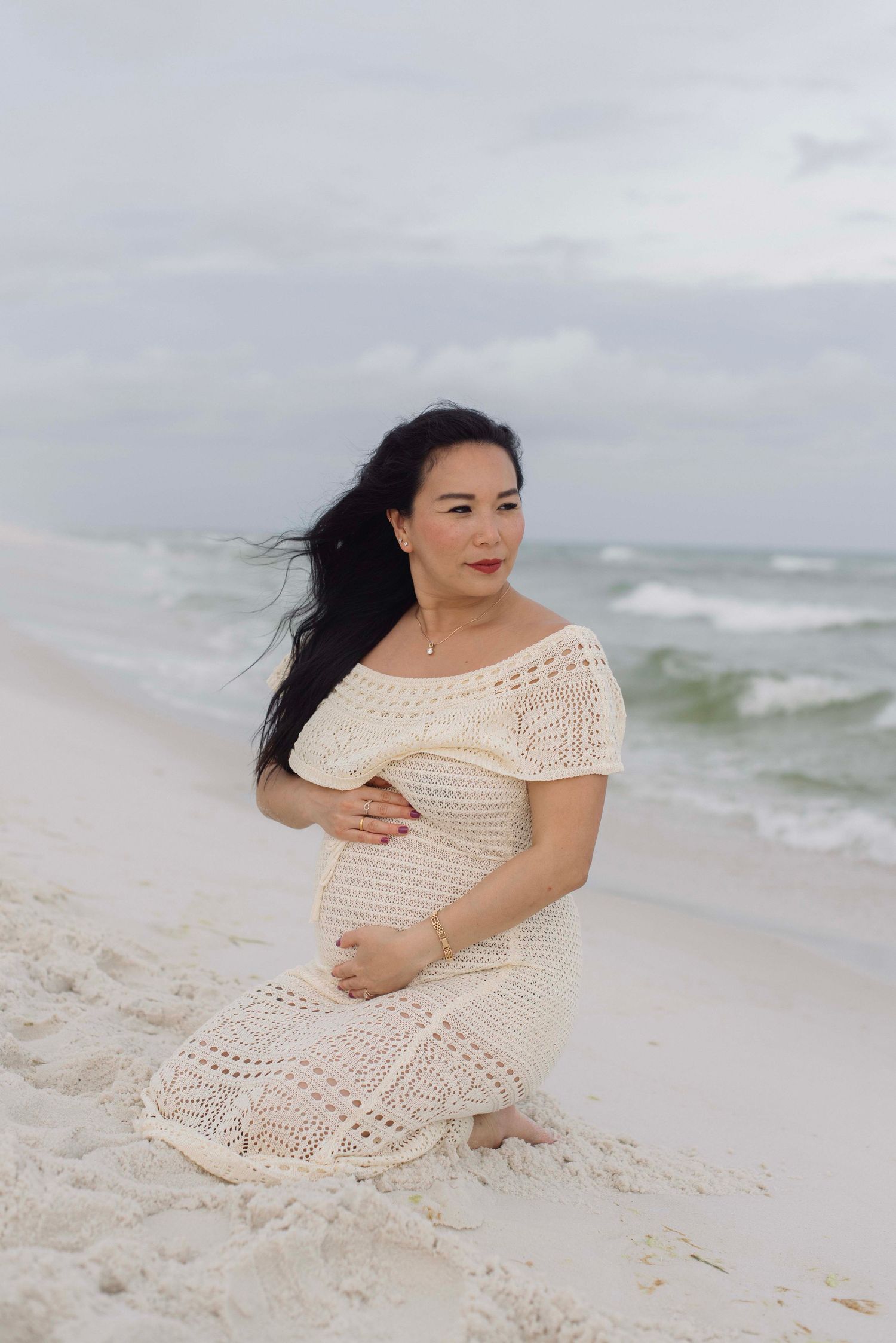 A person in a cream lace dress poses on a white sandy beach with ocean waves in the background.